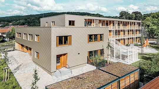 Aus erhöhter Perspektive zeigt das Bild den L-förmigen Neubau der Kindertagesstätte in Alzenau mit angrenzendem Wohntrakt – ein zukunftsweisendes Hybridgebäude in nachhaltiger Bauweise. Die vorgehängte PREFA Aluminiumfassade in Bronze mit Wandrauten 44 × 44 prägt die moderne, strukturierte Außenhülle des Kita-Bereichs. Großzügige Holzfenster und vertikale Lamellen schaffen ein freundliches, warmes Erscheinungsbild. Der rückwärtige Bereich mit Außentreppe, Spielflächen und Grünanlagen verdeutlicht die funktionale Trennung und gleichzeitige Verbindung der verschiedenen Nutzungen. Das begrünte Dach sowie die Materialkombination aus Aluminium, Holz und Beton spiegeln den hohen ökologischen Anspruch des Projekts wider – ein starkes Statement für nachhaltige Bildungs- und Wohnarchitektur.