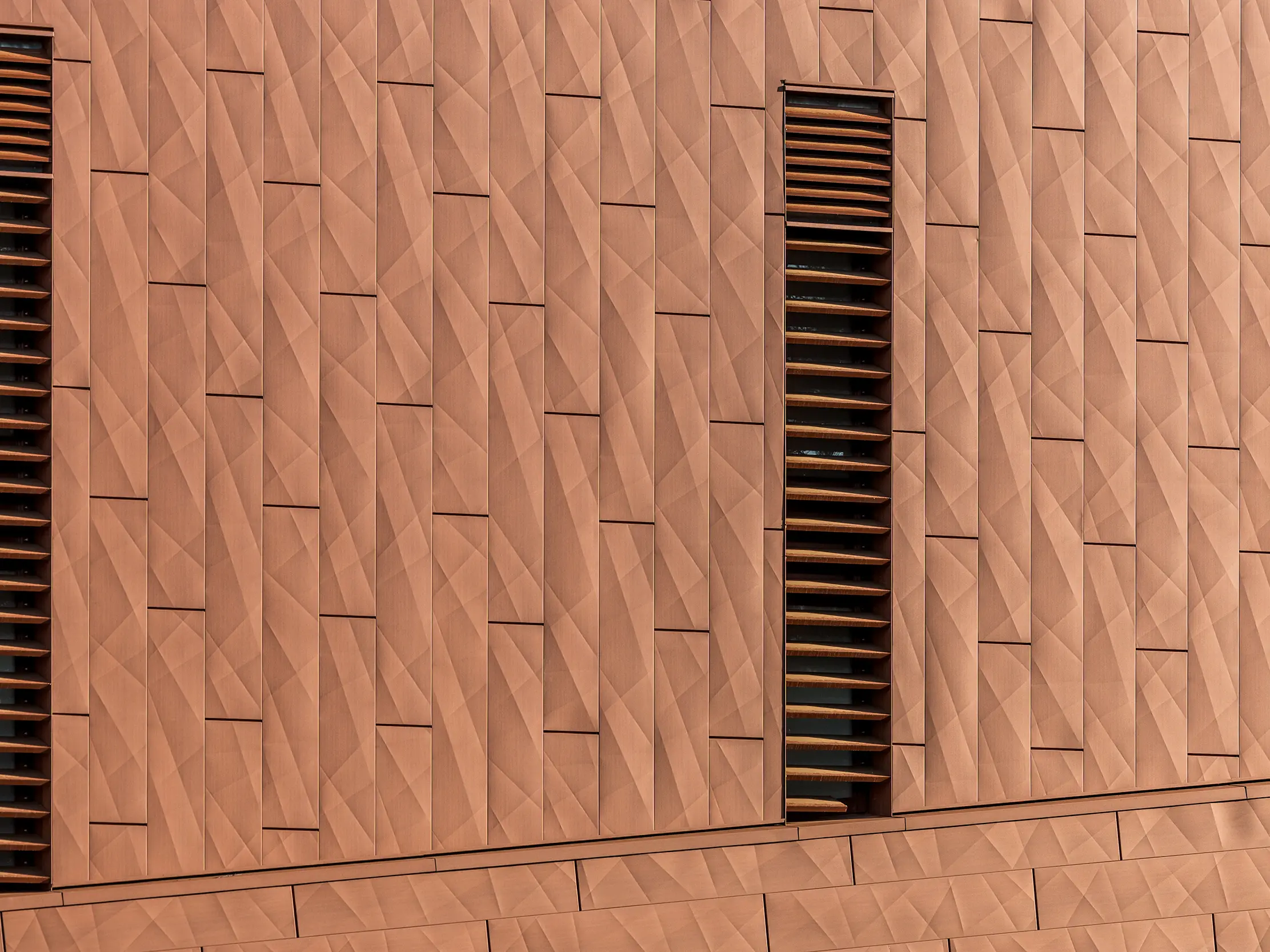 Detailaufnahme der PREFA Siding.X Aluminiumfassade am Trinkwasserpumpwerk WML in Lottum mit fein gefalteter, geometrischer Oberfläche in warmer Rostoptik. Die präzise angeordneten Paneele erzeugen ein lebendiges Spiel aus Licht und Schatten, während schlanke, vertikale Lamellenöffnungen als markante Eyecatcher das Fassadenbild strukturieren. Die leichten, langlebigen und vollständig recycelbaren PREFA Aluminium-Paneele überzeugen durch hohe Witterungsbeständigkeit, Farbkonstanz und eine nachhaltige, wartungsarme Gebäudehülle.