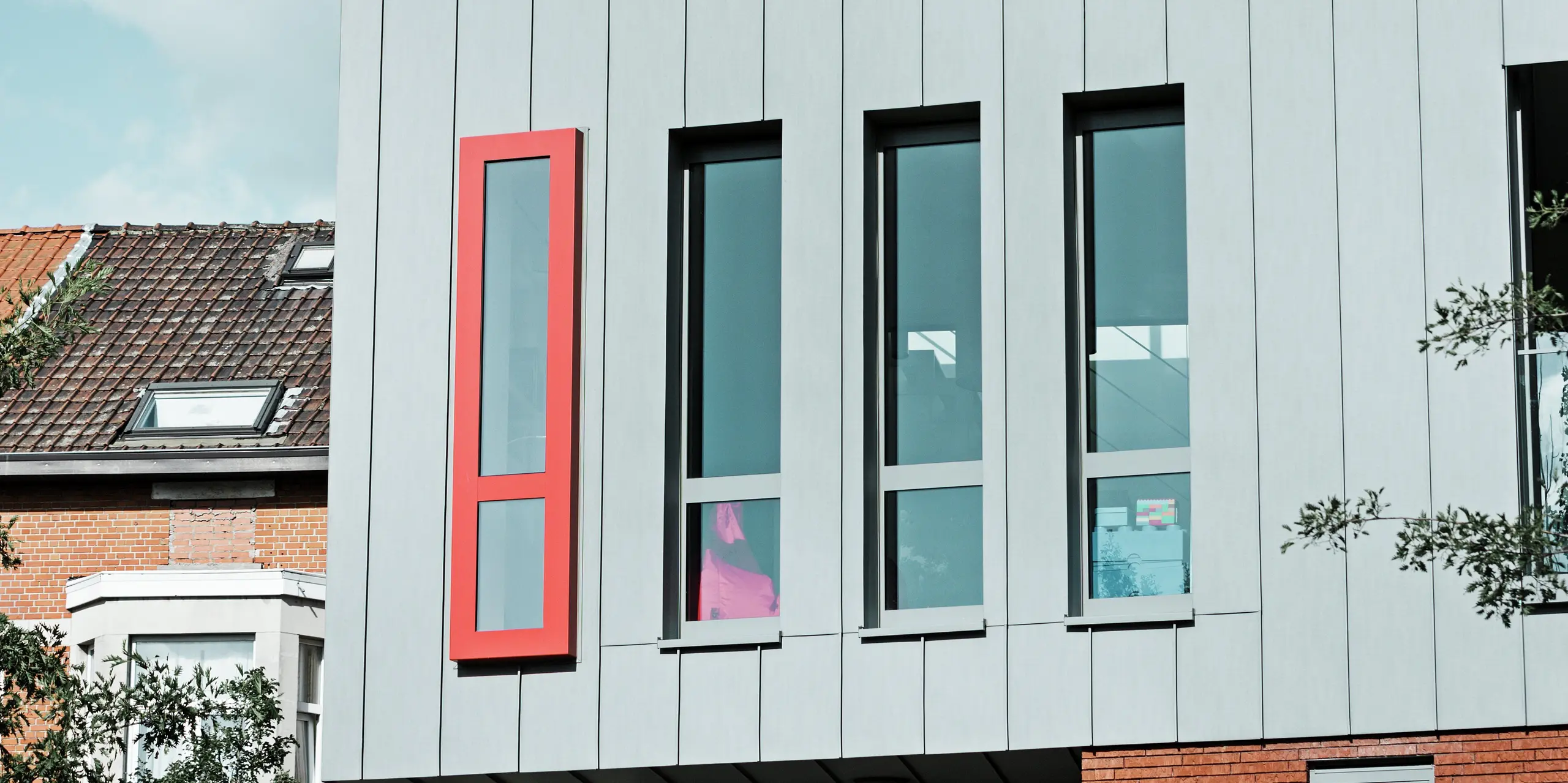 Diese Detailaufnahme zeigt die moderne PREFALZ-Aluminiumfassade in der Farbe Patinagrau mit vertikaler Stehfalzstruktur. Besonders ins Auge fällt das schmale, hochformatige Fenster mit dem kräftig roten Fensterrahmen, der einen architektonischen Akzent setzt. Die großzügigen Glasflächen sorgen für Licht im Gebäude. 