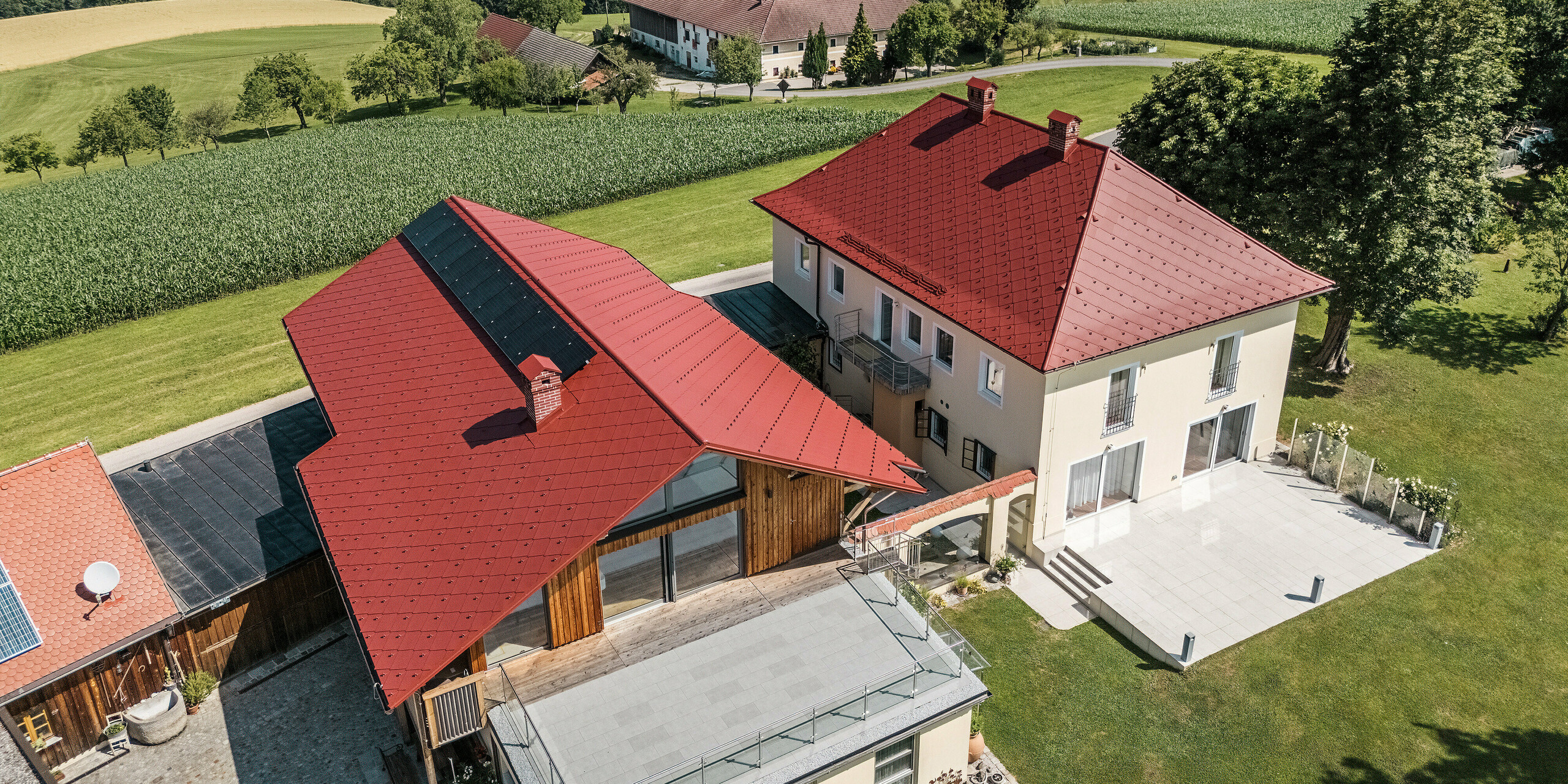 Die Luftaufnahme zeigt die sanierten Einfamilienhäuser in Aschach an der Steyr mit PREFA Dachrauten 44 x 44 in der Farbe P.10 Oxydrot. Die markante rote Dachfarbe harmoniert perfekt mit der Umgebung und unterstreicht die traditionelle Architektur der Häuser. Im Hintergrund ist die malerische Landschaft Oberösterreichs zu erkennen. Das Satteldach des im Vordergrund stehenden Hauses wurde mit einer kleinen Photovoltaikanlage ausgestattet.