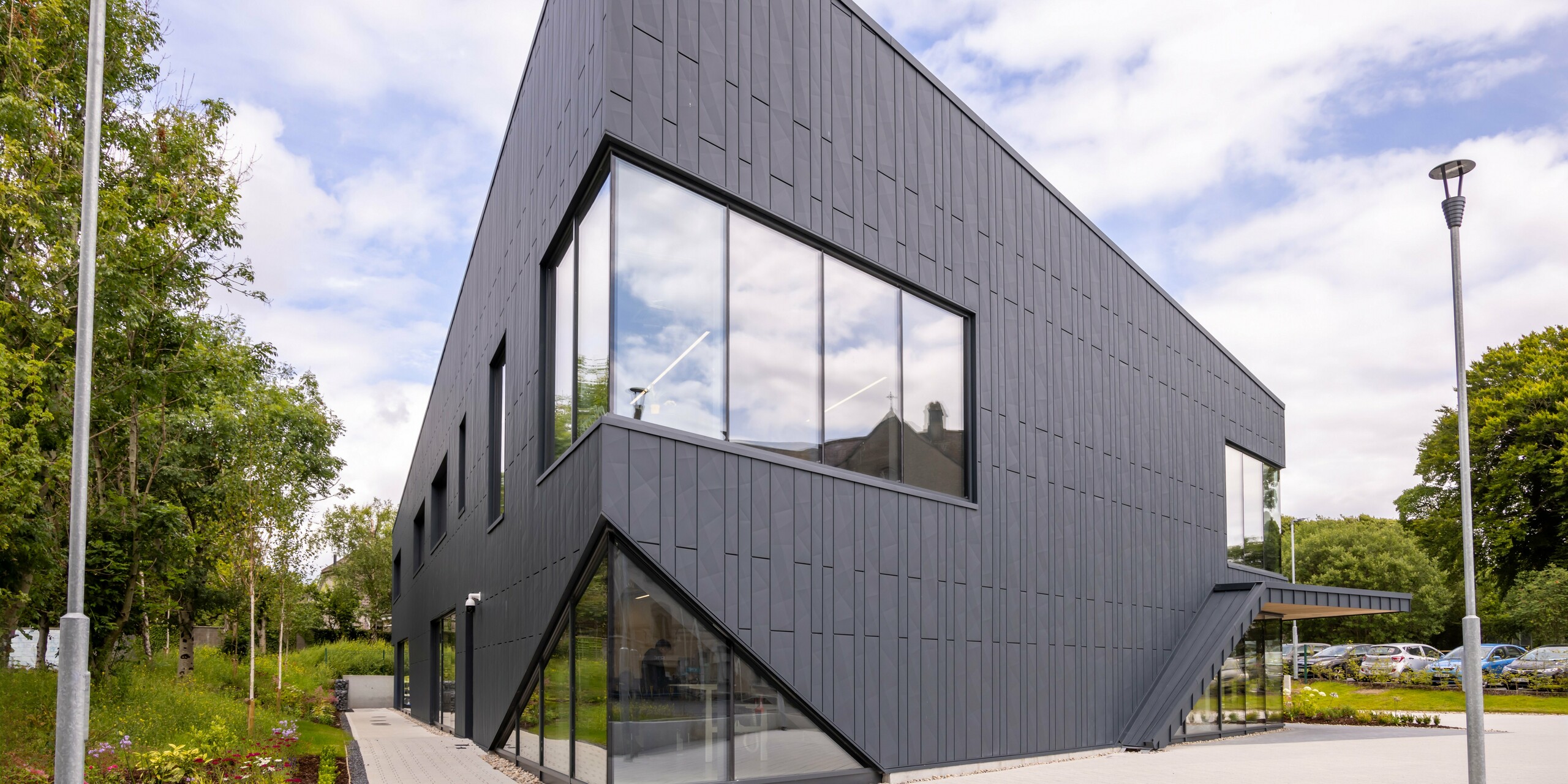 View of the front of the CREW building in Galway, Ireland, with a modern façade made of PREFA Siding.X façade panels and PREFALZ in the colour P.10 anthracite. The vertical lines of the façade and the large window areas emphasise the innovative and dynamic design of the building. The aluminium cladding blends harmoniously into the surrounding landscape and underlines the high-quality architecture of the building.