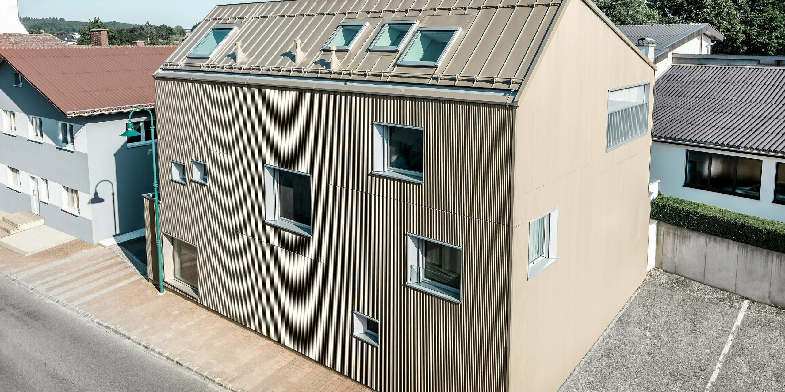 Vogelperspektive auf ein Einfamilienhaus in Frankenburg, Österreich, das hervorsticht durch seine PREFA Fassade in Form des Zackenprofils und einem PREFALZ-Dach, beide in abgestimmtem Bronze-Farbton. Die Fassade zeigt eine markante Textur, die das natürliche Licht einfängt und für ein lebendiges Schattenspiel sorgt. Mehrere Dachfenster lassen natürliches Licht ins Innere und unterstreichen die funktionale und ästhetische Integration moderner Architekturelemente. Das Haus fügt sich durch sein zeitgemäßes Design harmonisch in die typisch österreichische Dorf-Umgebung ein und setzt gleichzeitig einen kontrastreichen Akzent gegenüber den umliegenden traditionellen Gebäuden.