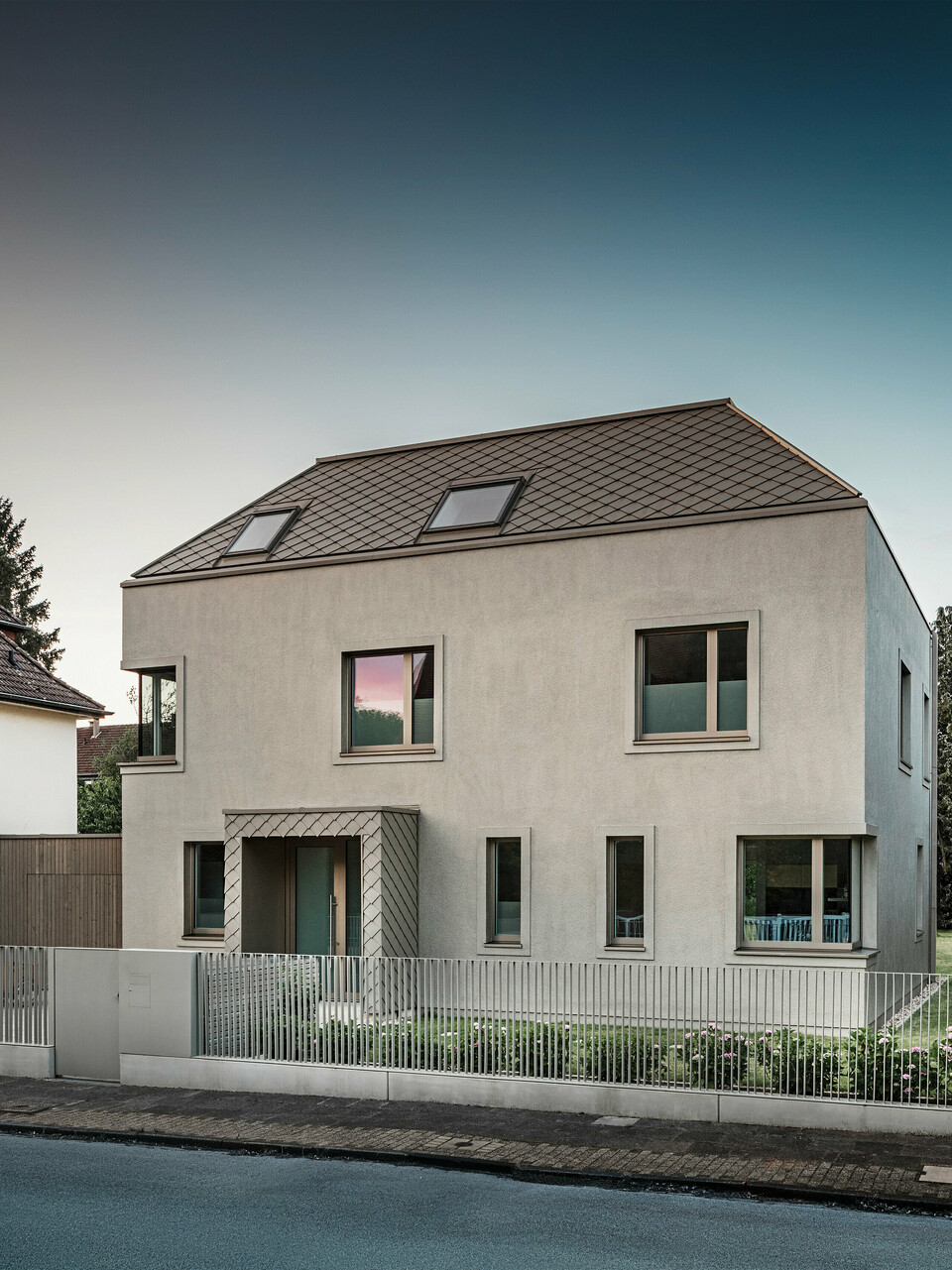 A modern detached house with a hipped roof and bronze-coloured aluminium roof and façade cladding from PREFA. The house is located in a quiet residential neighbourhood, surrounded by well-tended gardens and other detached houses. A low fence separating the property from the street can be seen in the foreground. The architecture of the house is characterised by clean lines and large windows that allow plenty of natural light into the interior.