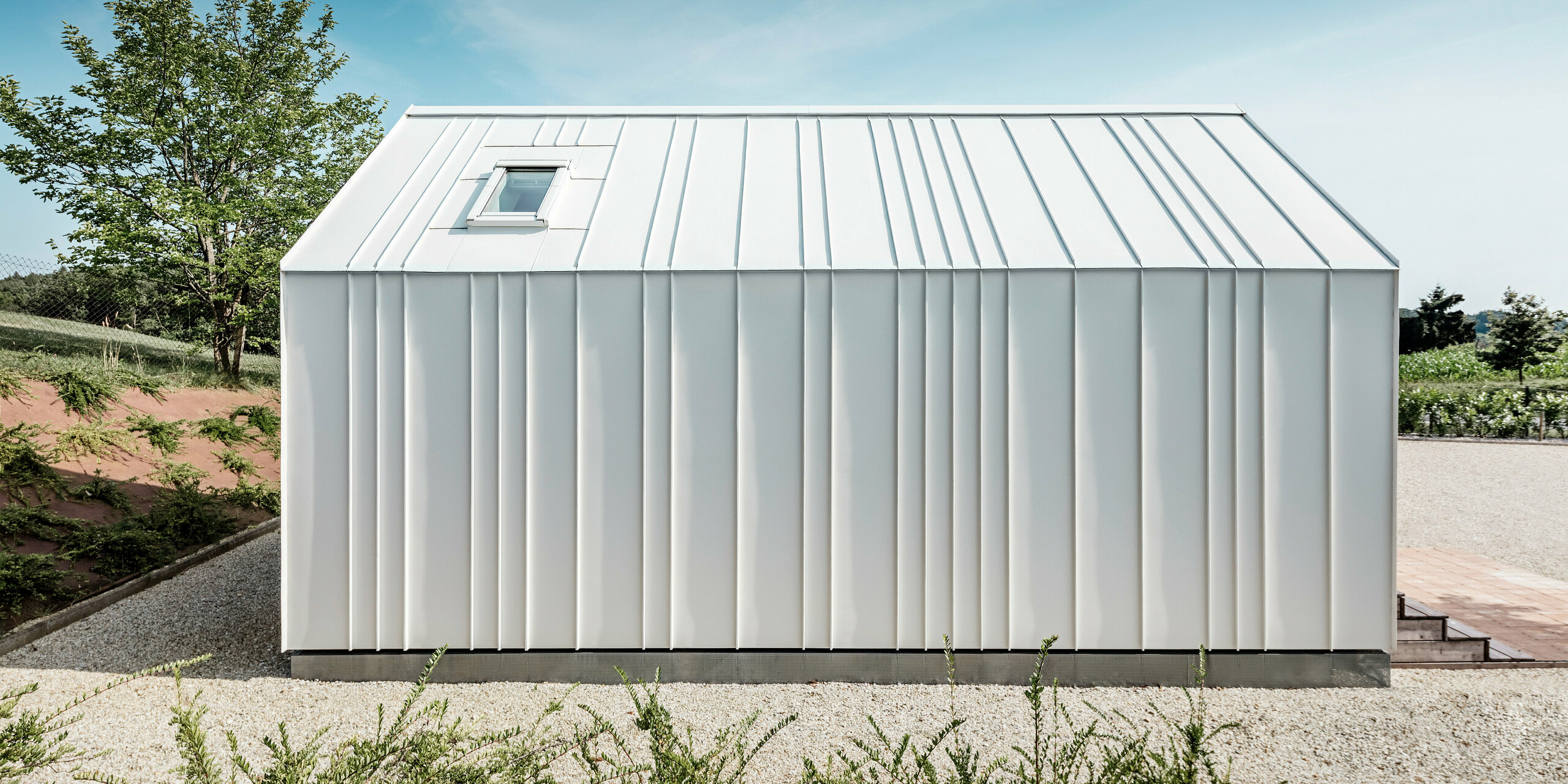 Side view of one of the tiny houses 'Pri Momi' in Kančevci, Slovenia. The micro house is encased in an elegant standing seam cladding made from the durable PREFALZ roof and façade system. The high-quality sheet metal cladding is finished in the colour P.10 pure white. The picture shows the precisely executed seams and the elegant lines of the white aluminium cladding, which give the building a modern and minimalist look. The natural surroundings and the clear, simple design harmonise perfectly with each other.