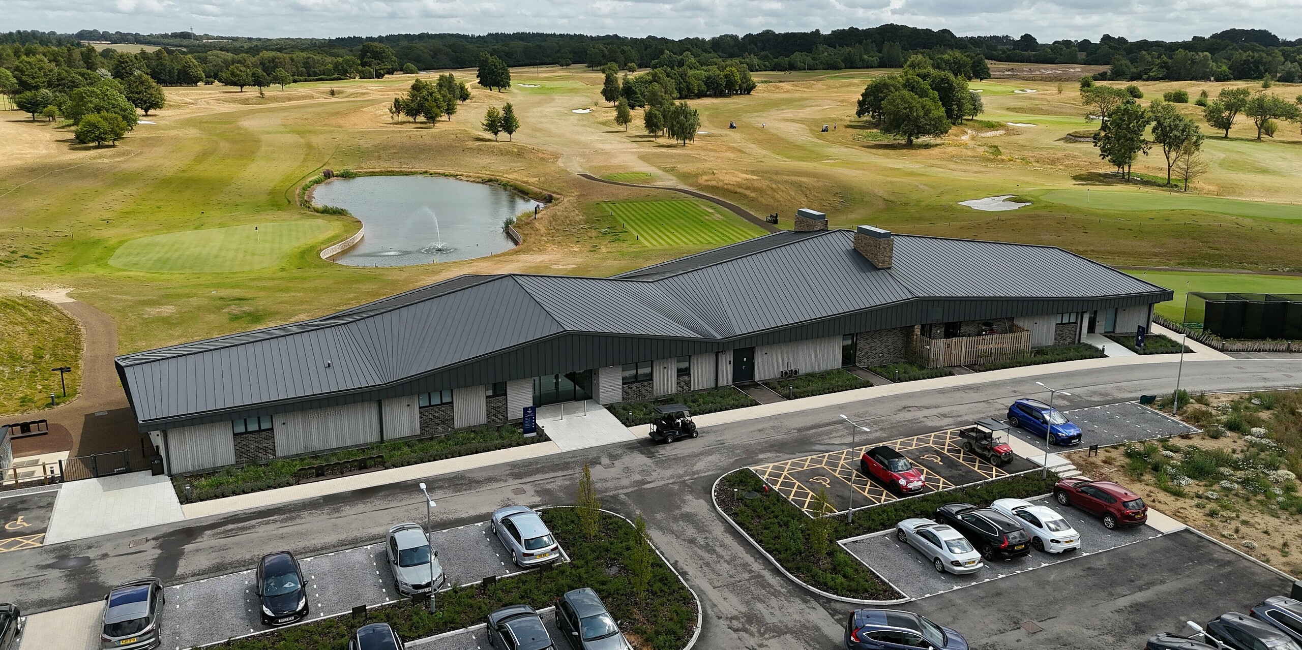 The modern new building at Basingstoke Golf Club in Hampshire impresses with its striking PREFALZ standing seam roof in P.10 dark grey. The dynamic roof shape and stylish façade made of wood and stone blend perfectly into the expansive golf course landscape. The building is picturesquely situated on the waterfront, surrounded by manicured green spaces. In the foreground is the car park with vehicles and a golf cart. The durable, low-maintenance aluminium of the roof provides long-lasting protection from the elements and gives the clubhouse a contemporary, functional aesthetic.