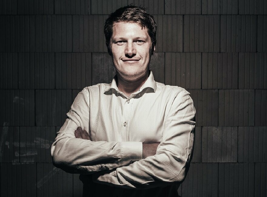 Portrait of the architect Andreas Gruber. He wears a white shirt and looks resolutely towards the camera, arms folded.