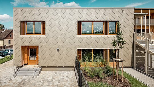 Die Rückseite der modernen Kindertagesstätte in Alzenau wird aus einer frontalen Perspektive gezeigt und beeindruckt mit klaren Linien und einer markanten Fassade aus bronzefarbenen PREFA Wandrauten 44 × 44. Die geometrische Fassadenstruktur erzeugt ein harmonisches Schuppenmuster, das durch die ruhige, horizontale Ausrichtung der Fenster akzentuiert wird. Großzügige Holzrahmen und vertikale Lamellen setzen natürliche Akzente und sorgen für eine warme, einladende Atmosphäre. Die durchdachte Kombination aus langlebigem Aluminium und regionalem Holz steht für nachhaltige Architektur mit hoher gestalterischer Qualität. Das Bild betont die präzise Ausführung, die hochwertige Materialwahl und die gelungene Integration des Gebäudes in das städtische Umfeld.