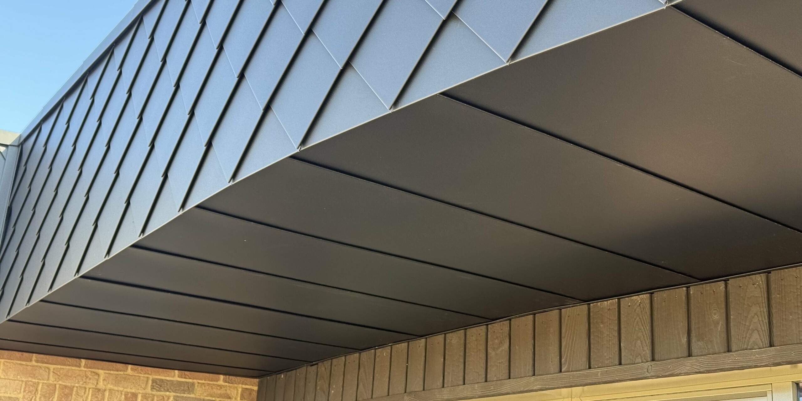 Detailansicht der modernen Fassadengestaltung eines Eco House im Projekt „Doone Brae Farm”. Im oberen Bereich der Fassade setzt die kleinformatige PREFA Wandraute in der Farbe P.10 Anthrazit markante Akzente. Die robusten Aluminiumrauten sind witterungsbeständig, langlebig und pflegeleicht – ideal für nachhaltige Architektur. Die Unterseite wird von glatten Aluminium-Paneelen ergänzt. Warme Klinkerflächen im unteren Fassadenbereich sorgen für einen spannenden Materialmix. Die Kombination aus Aluminium und Klinker vereint zeitlose Ästhetik mit höchster Funktionalität und macht das Gebäude zu einem echten Eyecatcher.