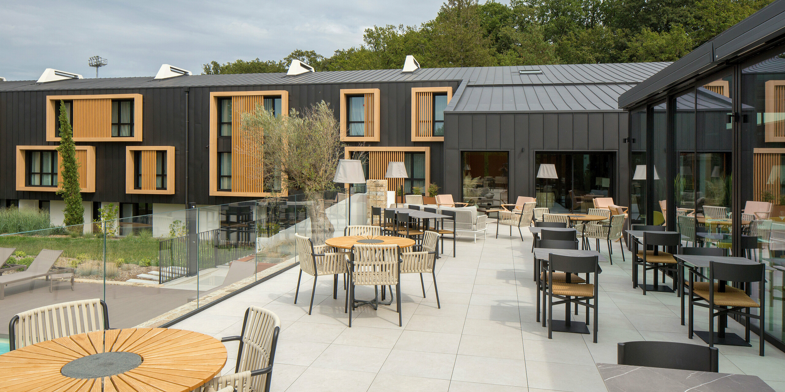 Terrassenbereich des Hôtel Parc du Landreau in Les Herbiers, Frankreich, mit Blick auf das schwarzgraue PREFALZ Dach- und Fassadensystem. Die naturbelassenen Holzrahmen um die Fenster ergänzen die witterungsbeständige Aluminiumverkleidung. Der Außenbereich bietet den Gästen eine einladende Atmosphäre mit bequemen Sitzgelegenheiten.