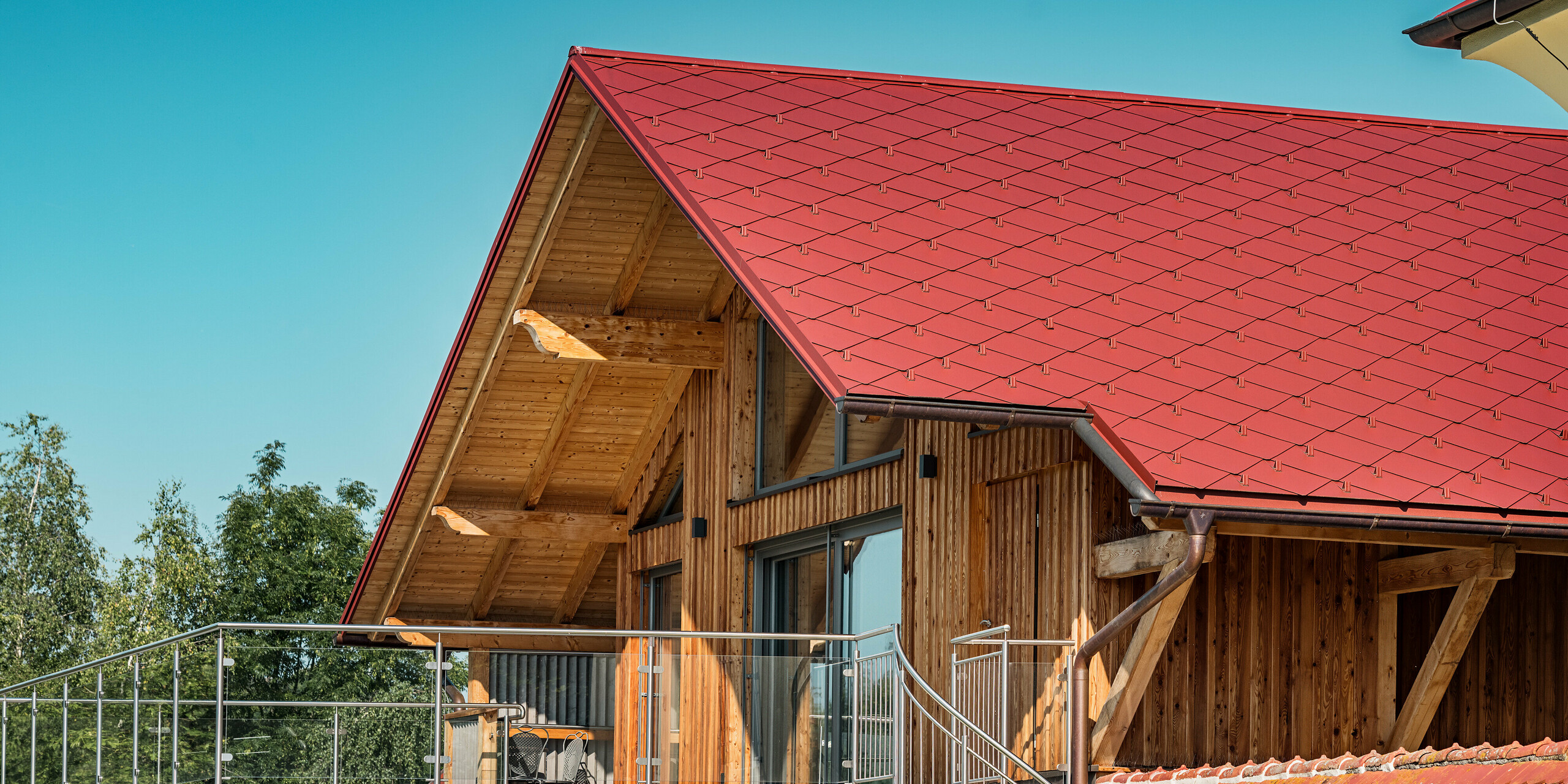 Das Bild zeigt einen detailreichen Ausschnitt des sanierten Satteldachs eines Einfamilienhauses in Aschach an der Steyr. Besonders auffällig ist die PREFA Dachraute 44x44 in der lebendigen Farbe P.10 Oxydrot, die das Dach ziert und dem Gebäude eine markante, aber harmonische Präsenz verleiht. Die rote Farbe ergänzt die stilvolle Holzfassade des Hauses perfekt.