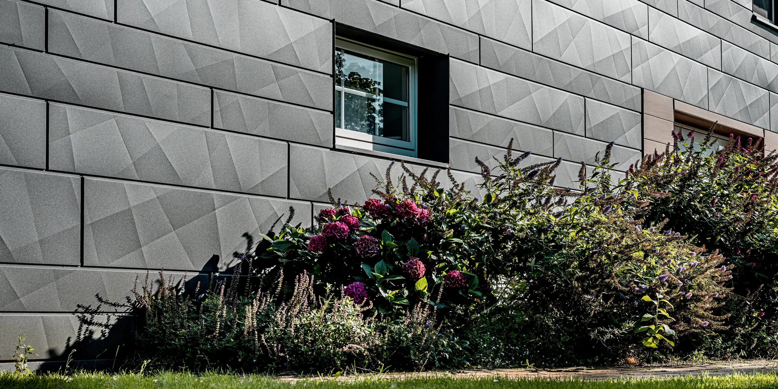 Nahaufnahme der sanierten Fassade eines Einfamilienhauses mit PREFA Siding.X in P.10 Dunkelgrau. Die großformatigen Aluminiumpaneele mit markanten Längs- und Querkantungen erzeugen eine lebendige, geometrische Oberflächenstruktur, die je nach Lichteinfall spannende Licht- und Schattenspiele zeigt. Ein präzise eingefasstes Fenster setzt einen klaren architektonischen Akzent und unterstreicht die hochwertige Verarbeitung. Im Vordergrund kontrastiert ein üppiges Blumenbeet mit farbigen Blüten und Grünpflanzen die kühle Eleganz der Aluminiumfassade. Die witterungsbeständigen, farbstabilen und pflegeleichten PREFA Aluminiumpaneele verbinden ausdrucksstarkes Design mit langlebiger Qualität und sind ideal für moderne, nachhaltige Fassadensanierungen.