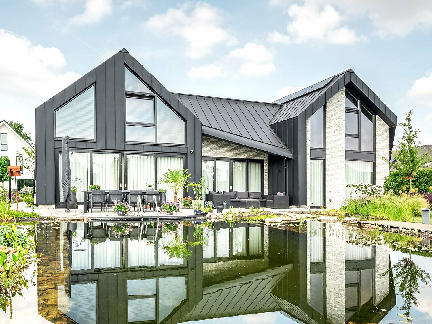 Modern detached house with characteristic, pointed gable roofs, covered with black Prefalz roof panels, reflected in a tranquil garden pond. The façade combines dark wall elements with light-coloured brick areas and large windows. A terrace with garden furniture invites you to linger and is harmoniously integrated into the lush, flowering garden landscape.