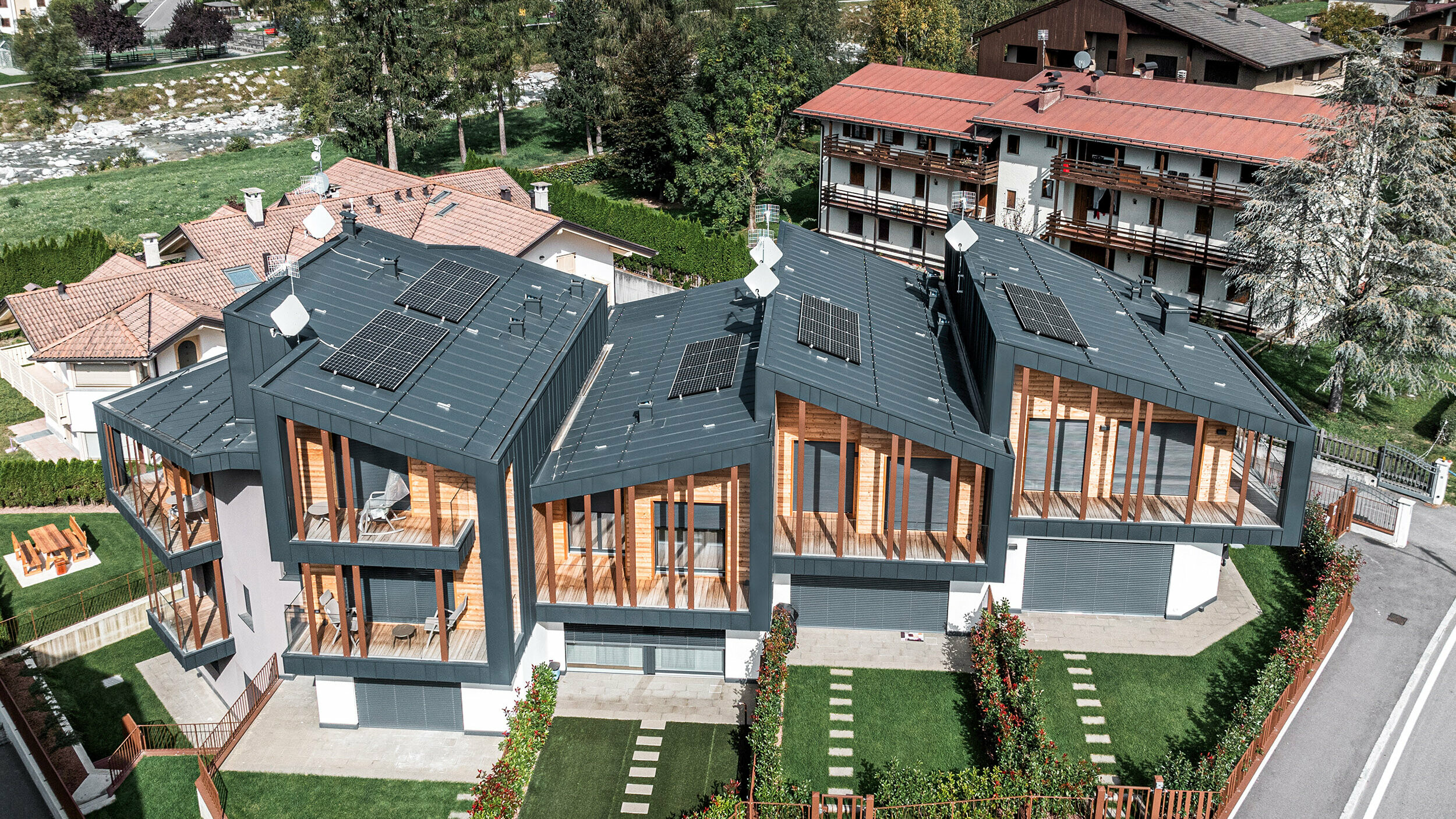 Luftaufnahme von modernen Ferienhäusern in den Dolomiten mit anthrazitfarbenen Prefalz-Metallpultdächern, die mit Solarmodulen ausgestattet sind. Die Fassaden zeigen eine Kombination aus dunkelgrauem Metall und natürlichen Holzelementen, harmonisch abgestimmt mit grünen Rasenflächen und verzierten Gartenbegrenzungen in einer Vorstadtgegend mit traditionellen Häusern und üppigen Bäumen im Hintergrund.