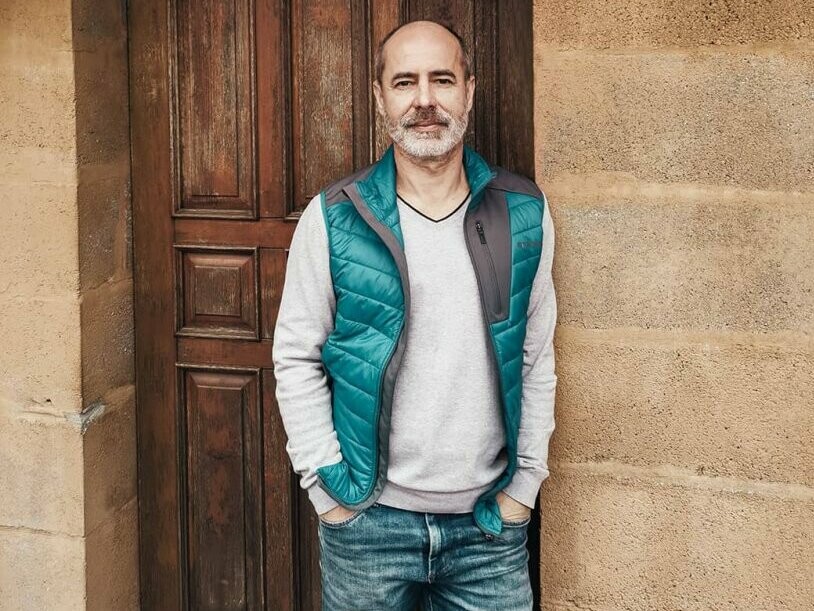 A picture of the architect Ferenc Kunis, standing in front of a brown door. His hands are tucked into his trouser pockets and he is looking into the camera.