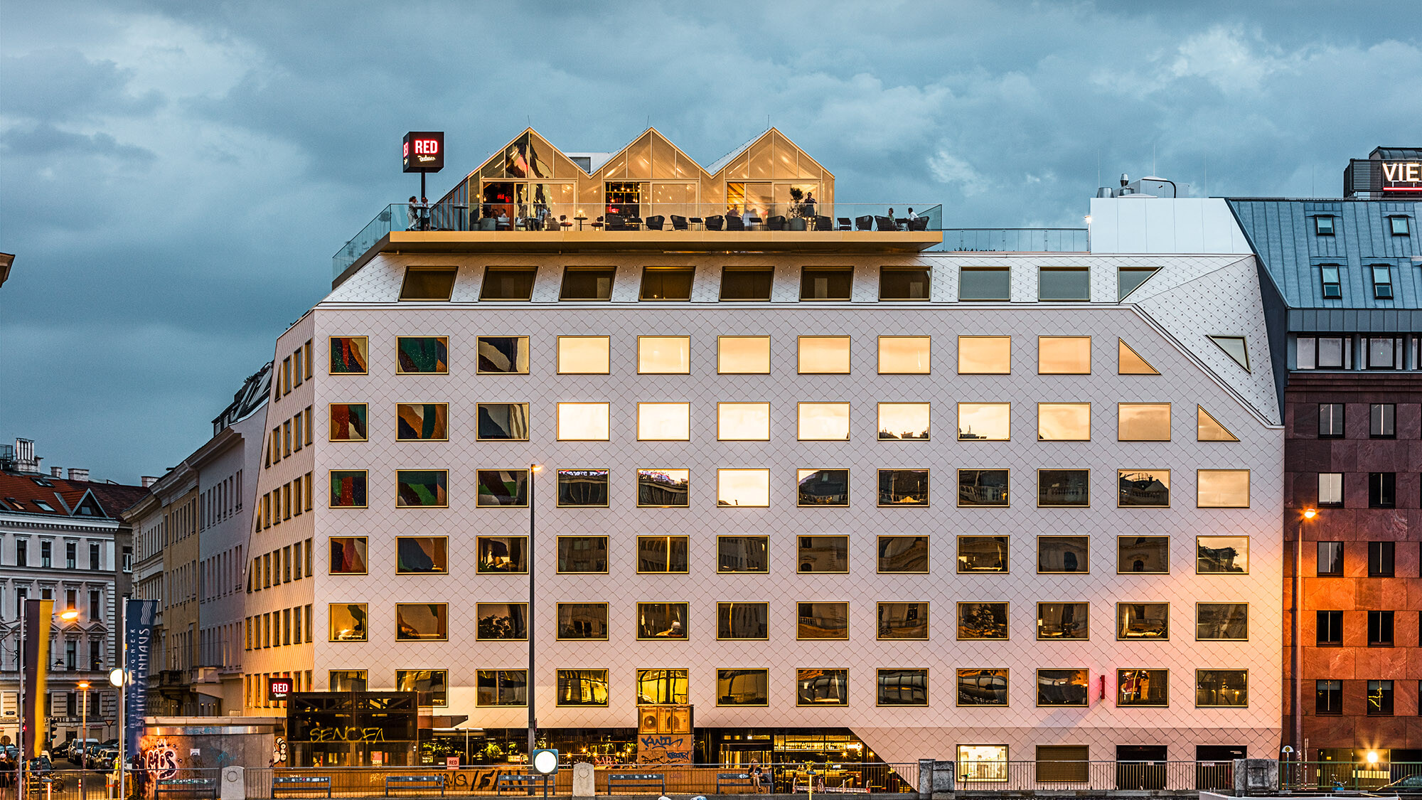 An evening shot of THE ROCK Radisson RED Vienna hotel with its lively surroundings; the buildings on the opposite side are mirrored in its windows.