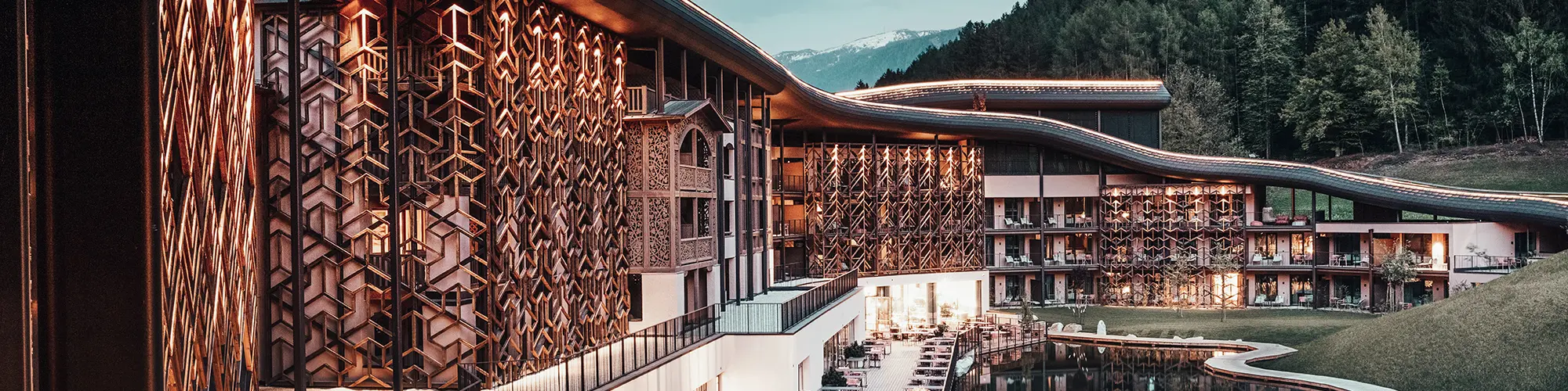 Evening shot, warm light illuminates the wooden façade and the aluminium roof, below the terrace with seating, in front of the lake, the green forest in the background