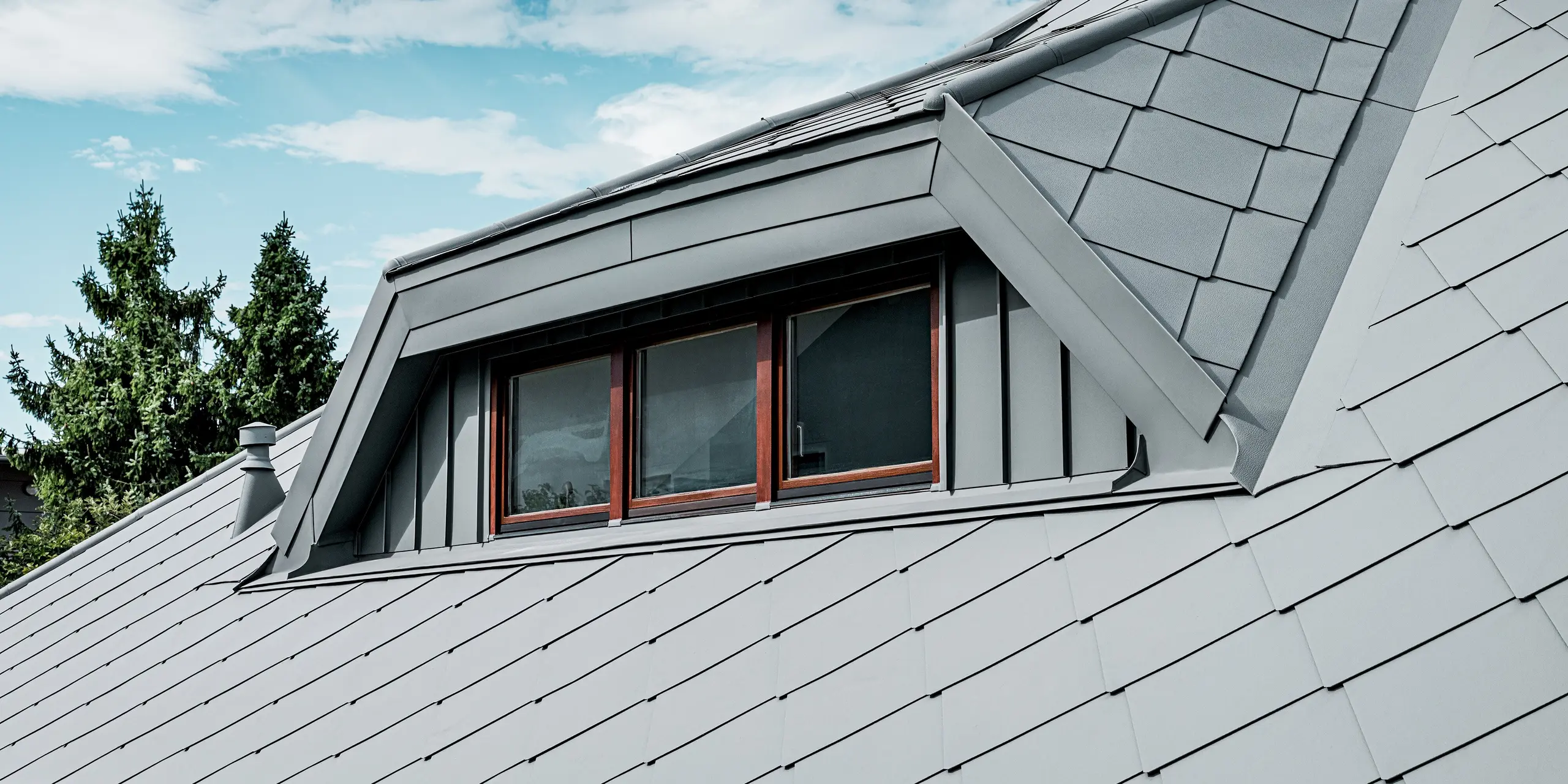Das Bild zeigt eine Nahaufnahme der markanten Trapezgaube eines Einfamilienhauses, deren Oberfläche vollständig mit PREFA Aluminium in P.10 Hellgrau gestaltet ist. Die präzise integrierte Dachraute 44 × 44 und die klar definierten Kanten der Gaubenkonstruktion erzeugen eine moderne, hochwertige Optik. Die feine Stucco-Struktur verleiht dem Aluminiumdach zusätzliche Tiefe und Robustheit. Unter dem hellen Himmel wirkt das langlebige, witterungsbeständige PREFA Dachsystem besonders elegant und stabil.