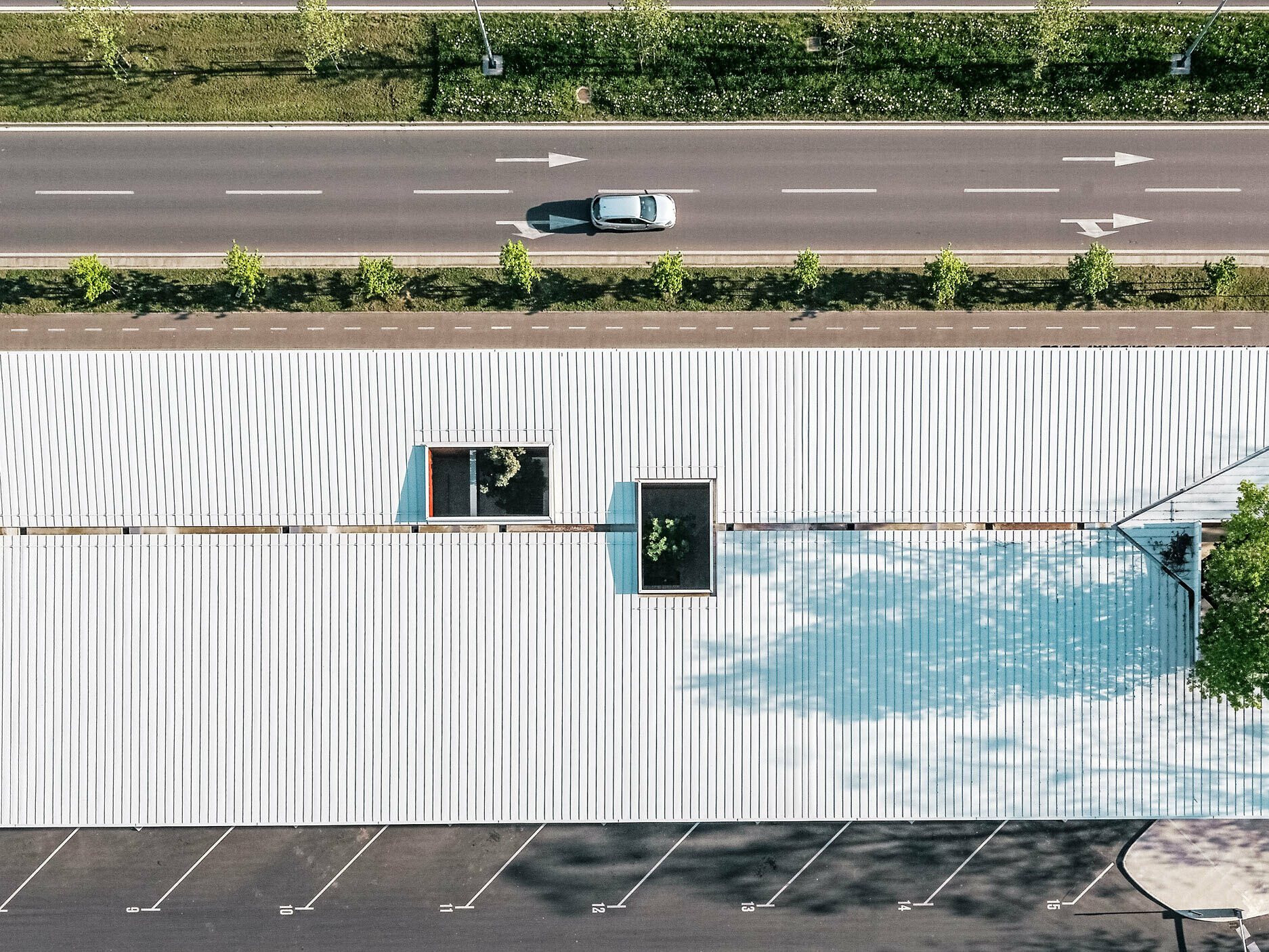 The picture shows an aerial view of the bus stop in Croatia with a white Prefalz roof from PREFA. The long building has several rectangular skylights arranged in different positions, allowing the green foliage of trees to show through. A small car park can be seen in the foreground, while a two-lane road with a line of trees runs along in the background. 