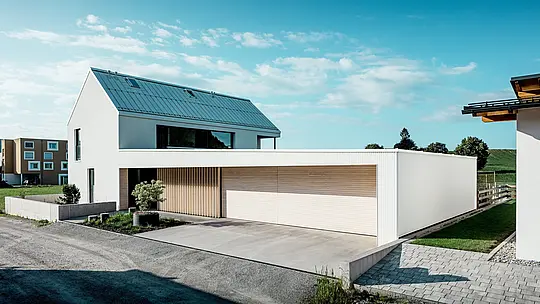 Das Bild zeigt das moderne Einfamilienhaus in Freistadt, das sich durch seine schlichte, geometrische Form und die Kombination von reinweißer PREFALZ-Aluminium-Fassade und Holzvertäfelungen auszeichnet. Die Holzverkleidung am Garagentor bietet einen warmen, natürlichen Kontrast und fügt sich perfekt in das moderne Design ein. Das Dach, ebenfalls mit PREFALZ gedeckt, sorgt für eine nahtlose, wetterfeste Oberfläche. Diese Kombination aus pflegeleichtem Aluminium und Holz schafft eine ästhetisch ansprechende und funktionale Lösung für nachhaltige Architektur. Der weitläufige, grüne Außenbereich und der klare Himmel im Hintergrund unterstreichen die moderne Lebensqualität, die dieses Haus bietet.