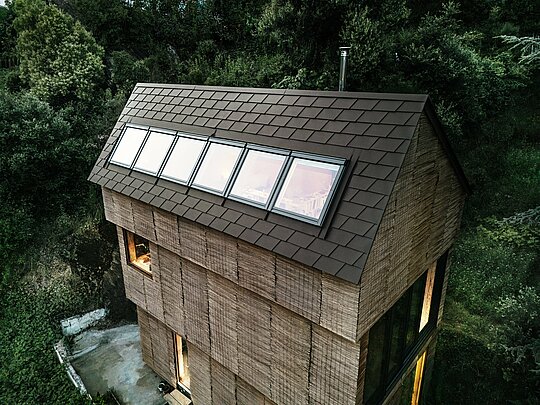 Dachansicht eines Hauses mit PREFA Dachschindeln in P.10 Nussbraun und integrierten Dachfenstern, umgeben von natürlicher Landschaft im Westen Frankreichs.