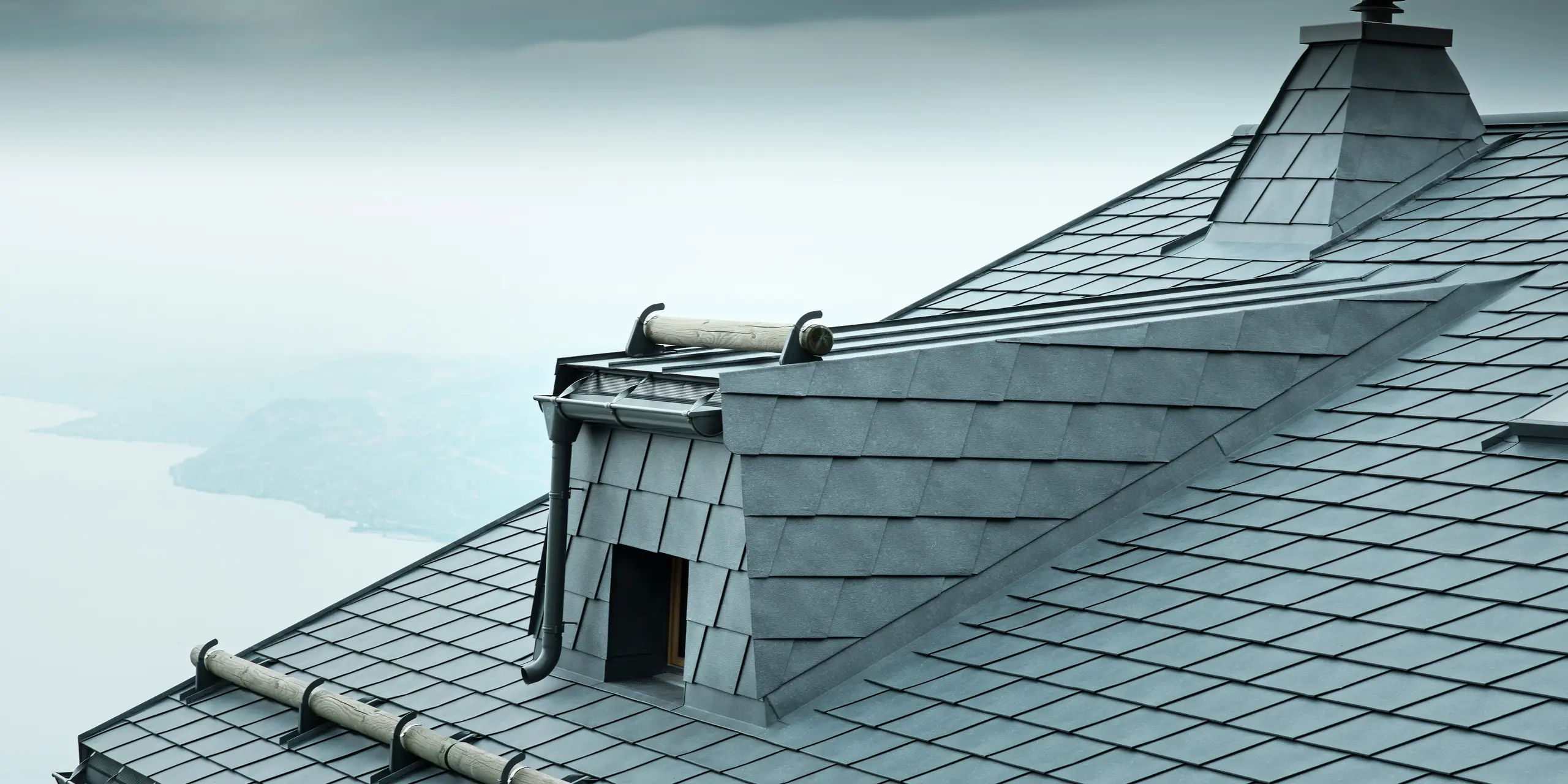 Die Dachansicht dieser modernen Holz-Lodge in der Schweiz zeigt steingraue PREFA Aluminium-Dachschindeln und eine große Dachgaube. Das langlebige Aluminiumdach schützt das Gebäude vor den extremen Wetterbedingungen am Genfersee. Der raffinierte Schneefang aus Rundholz und der markante Kamin betonen das einzigartige Design der Architektur.