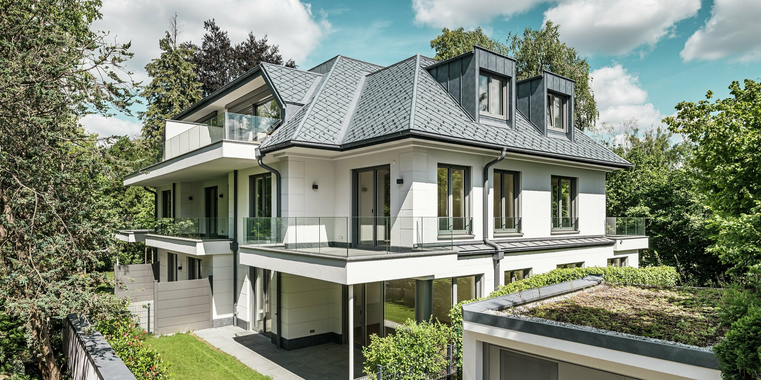Ansicht eines modernen Mehrfamilienhauses in der Nähe von München mit stilvollem Design. Das Dach wurde mit PREFA Dachrauten 29 × 29 in P.10 Steingrau gedeckt. Die markante Dachkonstruktion wird durch elegante Flachdachgauben aus PREFALZ akzentuiert, die sowohl ästhetisch als auch funktional überzeugen. Der gepflegte Gartenbereich sowie die großzügige Terrasse betonen die hochwertige Architektur und schaffen eine harmonische Verbindung zwischen Innen- und Außenbereich. Die langlebigen und witterungsbeständigen PREFA-Produkte garantieren eine perfekte Integration in die grüne Umgebung und bieten dauerhaft Schutz und Schönheit.