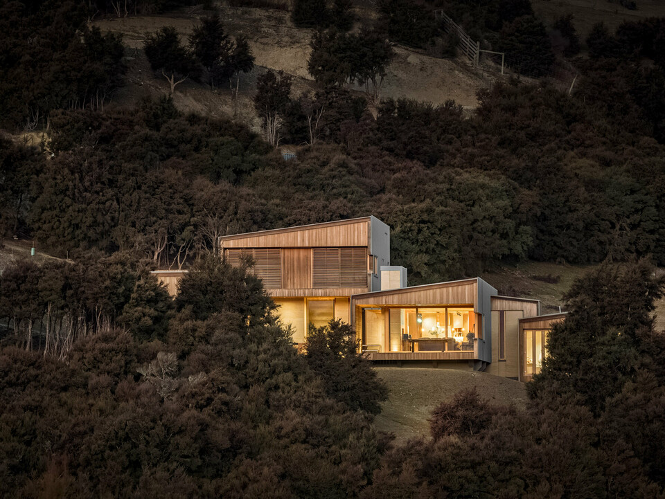 Evening shot of the detached house, illuminated from inside and embedded into the hilly landscape in the area around Wanaka, New Zealand. Part of the building is covered by the bush forest.