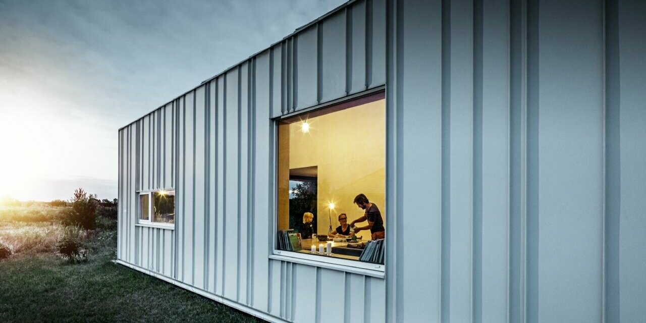 Angled standing seam façade with PREFALZ in different panel widths in P.10 Prefa white photographed at dusk. Across the dining room, you can make out a family having dinner. 