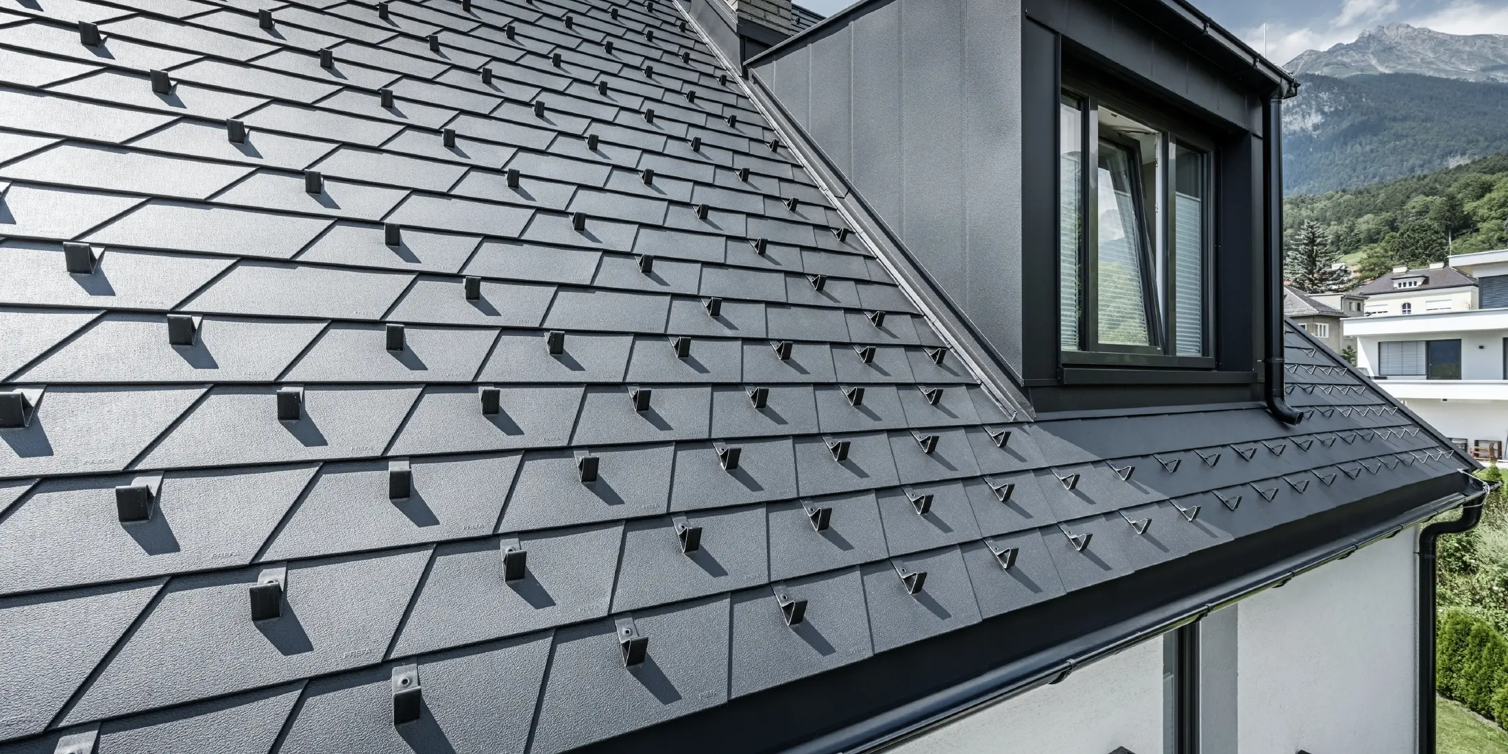 Detailed view of a roof with dormer and window, PREFA roof shingles in black with snow guards are laid on the roof surface.