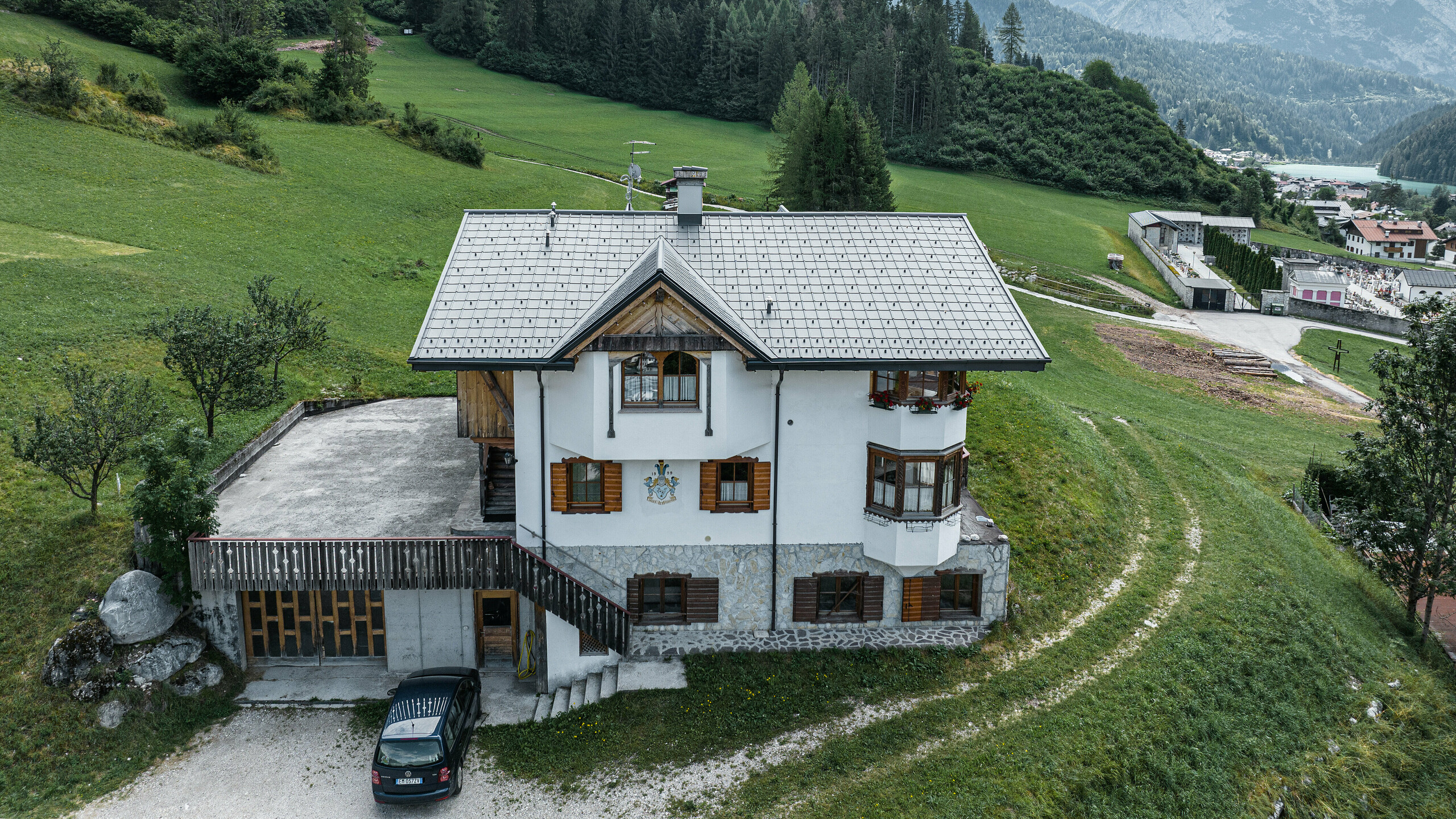 A bird's eye view of the entrance to the traditional mountain hut in Auronzo di Cadore after the roof renovation with PREFA tiles made of aluminium in P.10 light grey. The building, with its characteristic Alpine charm and modern sheet metal roof, blends harmoniously into the lush green landscape and the impressive mountain panorama in the background, demonstrating a successful combination of functionality and regional architecture.