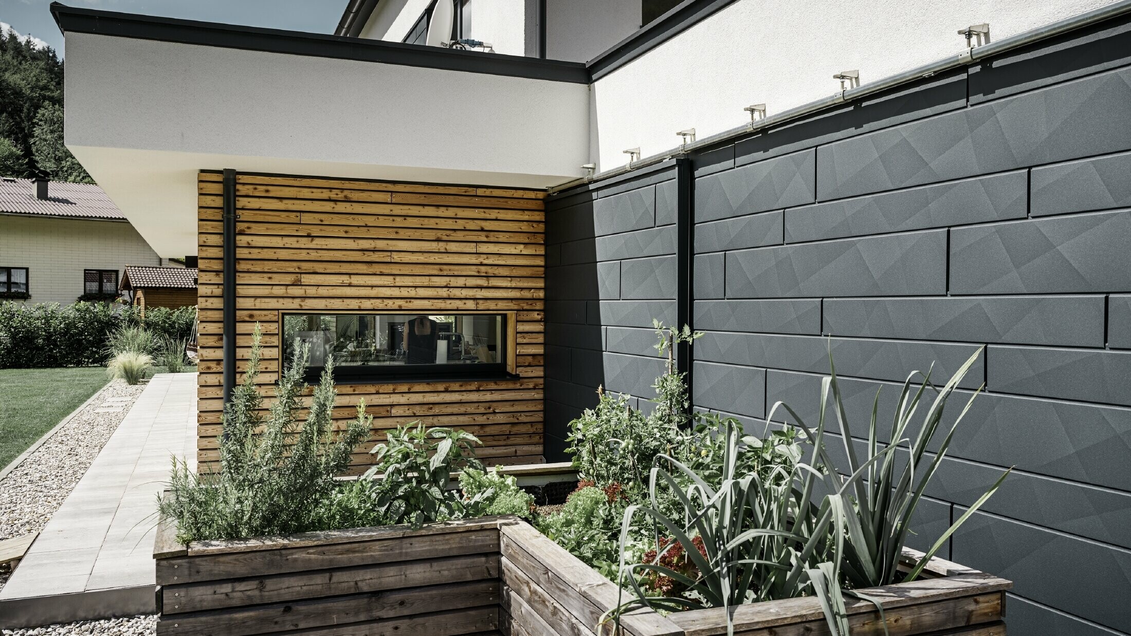 This image shows a close-up of the detached house, focusing on the façade cladding made from PREFA Siding.X in P.10 anthracite. The dark façade panels, with their distinctive crinkled finish, provide an appealing visual contrast to the vibrant, timber-clad areas and the surrounding green spaces. Raised beds, planted with various plants and shrubs, are positioned in front of the aluminium façade, emphasising the house’s natural and sustainable character. The combination of modern materials and integration into the garden area conveys an image of contemporary architecture that blends harmoniously into its surroundings.