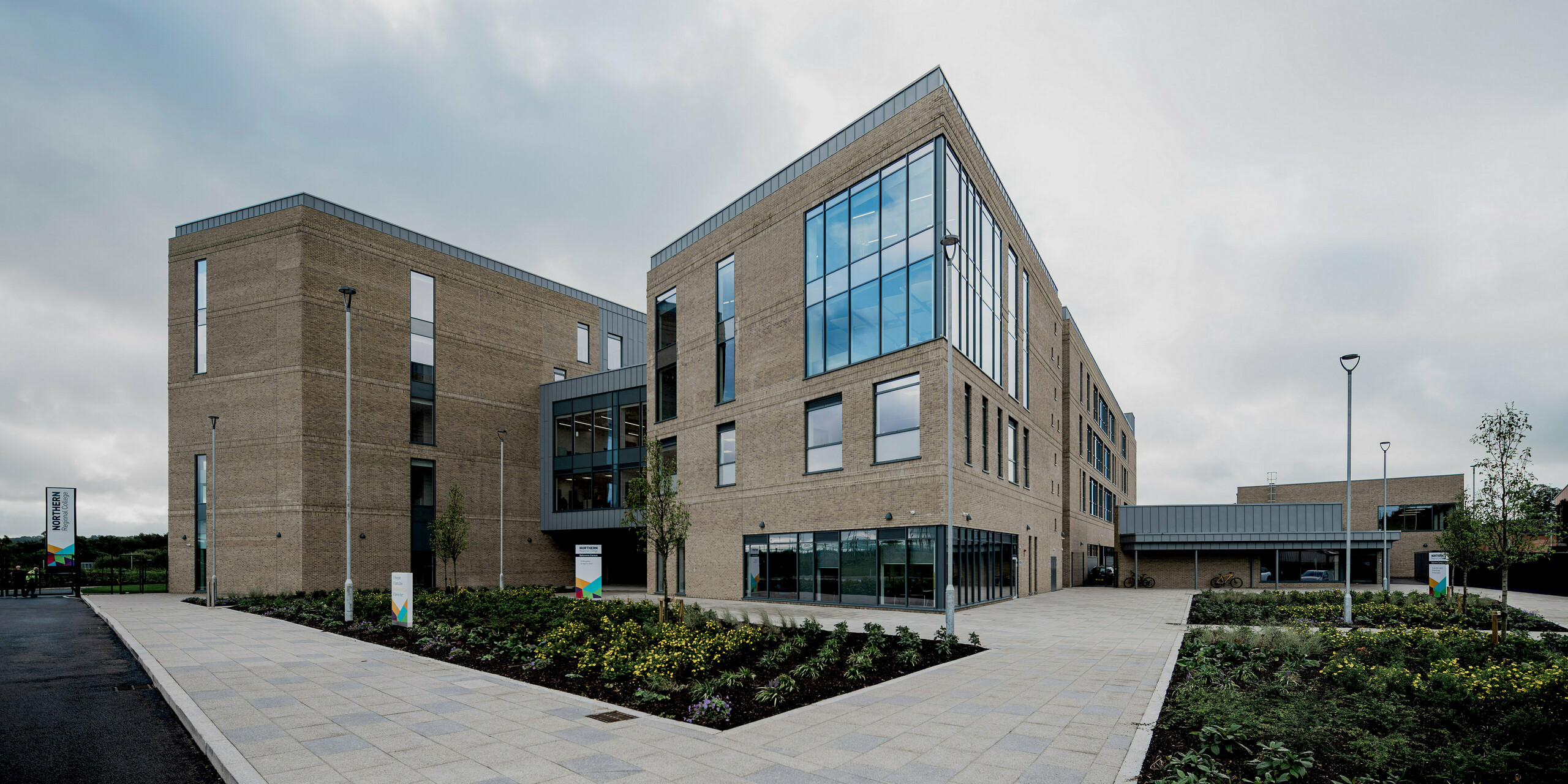 A wide view of the Northern Regional College in Ballymena reveals a modern campus composed of clearly structured brick volumes connected by glazed sections and open pedestrian paths. Along the rooflines and façade edges, PREFALZ standing seam cladding in patina grey create crisp horizontal accents. The refined metal surfaces contrast with the textured brickwork and open sky, enhancing the architectural clarity. PREFA aluminium offers durability, low maintenance and a timeless aesthetic ideal for contemporary educational buildings.