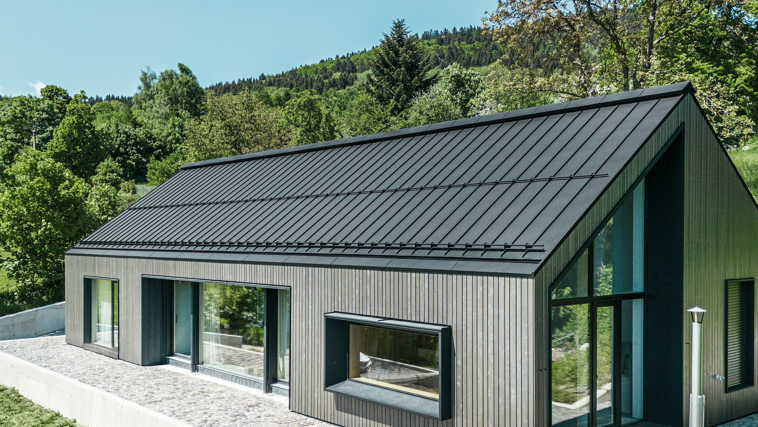 Ein modernes Holzmassivhaus in Kuncice (CZ) wurde mit großen Fensterflächen ausgestattet. Das dunkle Holzmassivhaus verfügt über ein Dachsystem aus PREFALZ in der Farbe P. 10 Schwarz. Im hinteren Außenbereich steht ein Whirlpool mit einer Holzverkleidung.