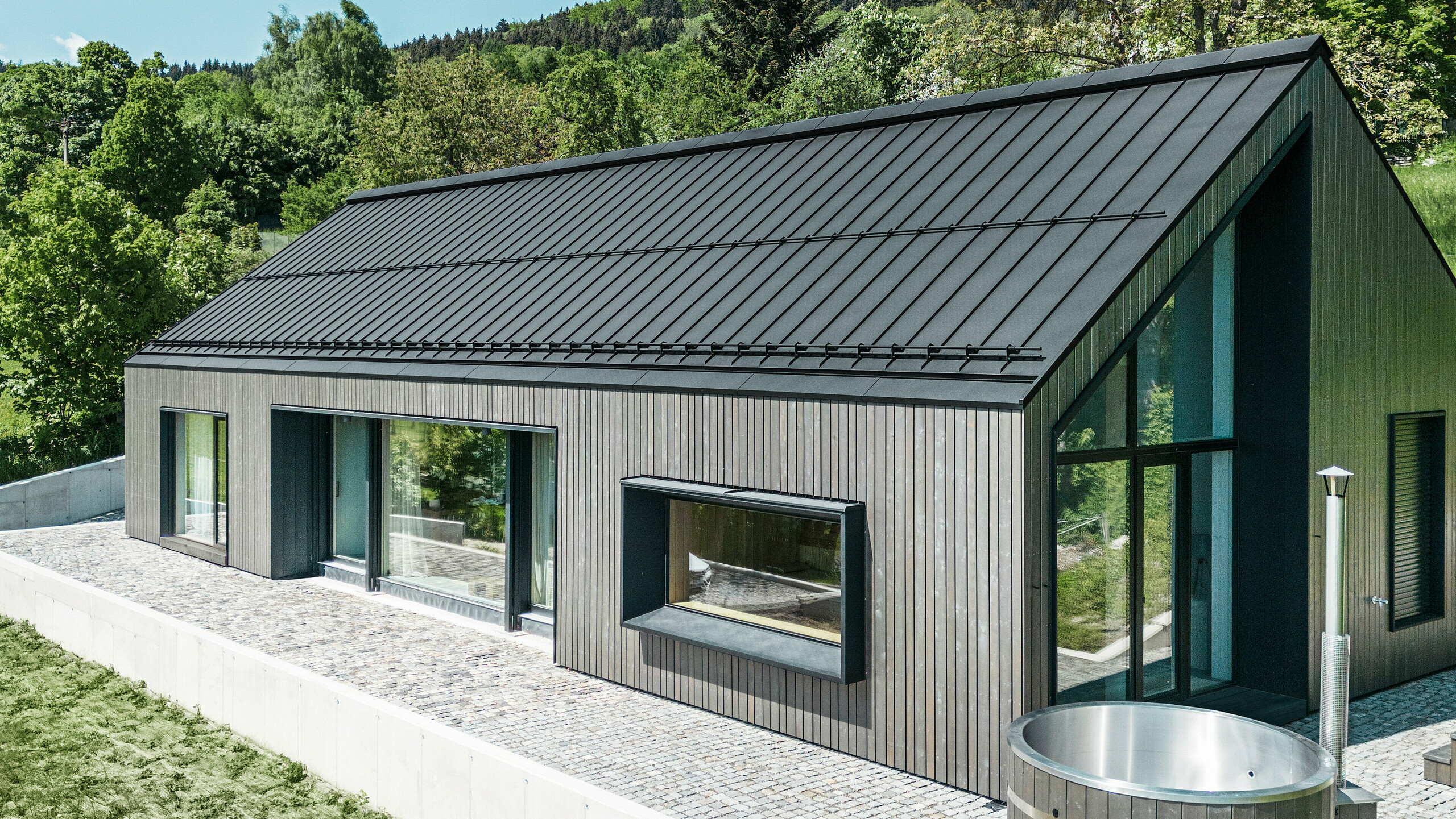 A modern wooden house in Kuncice (CZ) was equipped with large windows. The dark wooden house has a PREFALZ roof system in the colour P.10 black. In the rear outdoor area, there is a hot tub with wooden panelling.
