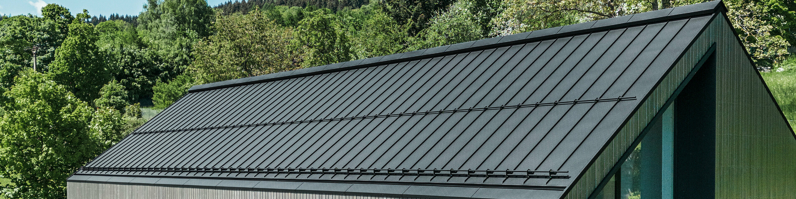 A modern wooden house in Kuncice (CZ) was equipped with large windows. The dark wooden house has a PREFALZ (standing seam) roof system in the colour P.10 black. In the rear outdoor area, there is a hot tub with wooden panelling.
