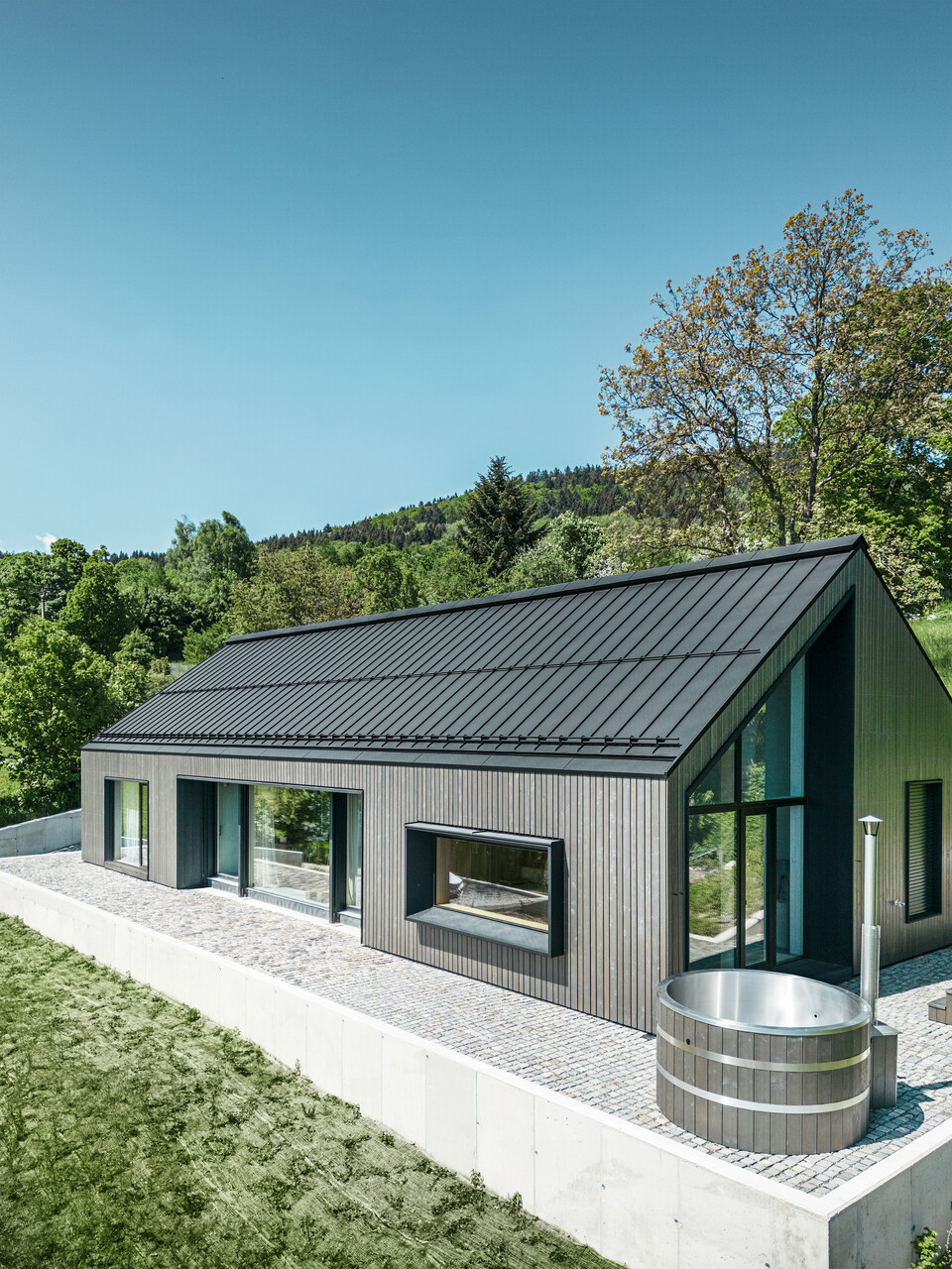 A modern wooden house in Kuncice (CZ) was equipped with large windows. The dark wooden house has a PREFALZ roof system in the colour P.10 black. In the rear outdoor area, there is a hot tub with wooden panelling.