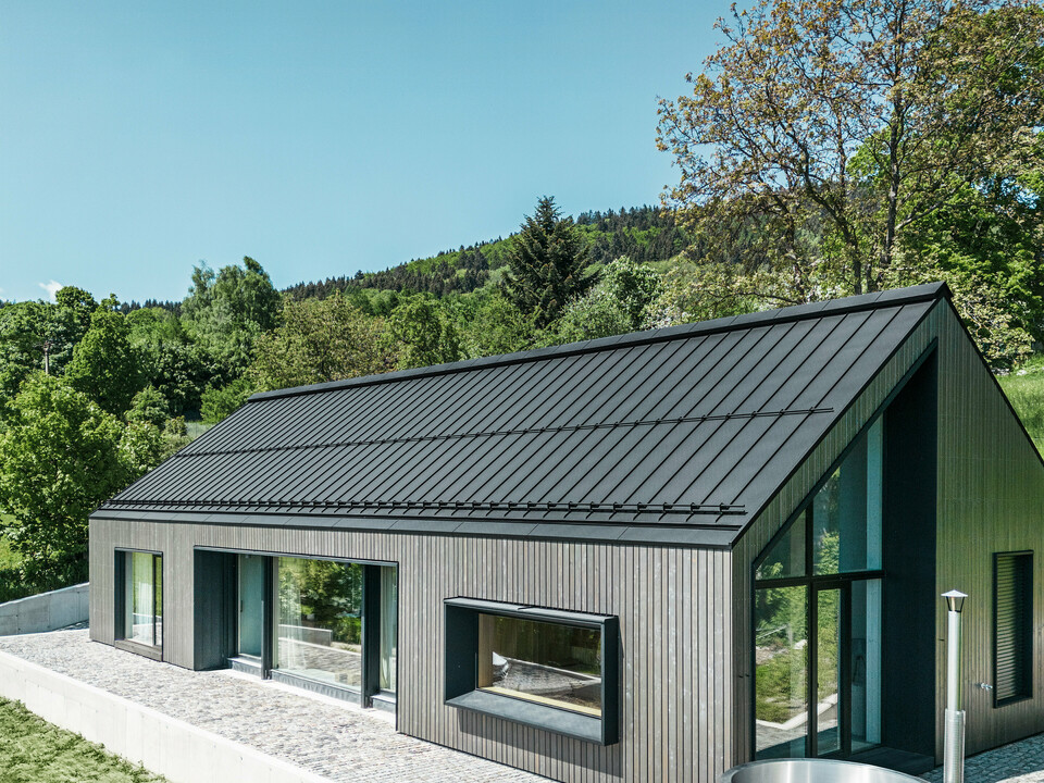 A modern wooden house in Kuncice (CZ) was equipped with large windows. The dark wooden house has a PREFALZ (standing seam) roof system in the colour P.10 black. In the rear outdoor area, there is a hot tub with wooden panelling.