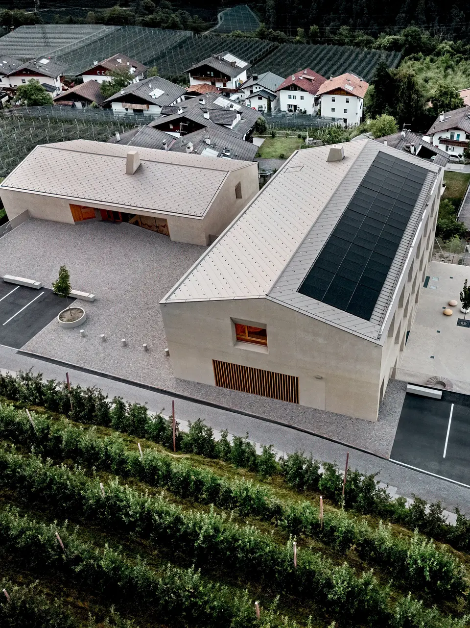 Eine Luftaufnahme des Bildungszentrums Riffian in Südtirol mit Kindergarten, Grundschule und Sportplatz. PREFA Dachrauten 44 × 44 in der Farbe P.10 Bronze bilden ein präzises und langlebiges Aluminiumdach, das sich harmonisch in die alpine Landschaft einfügt. Die moderne Architektur zeichnet sich durch ihre Witterungsbeständigkeit und ihr klares Design bis ins kleinste Detail aus.