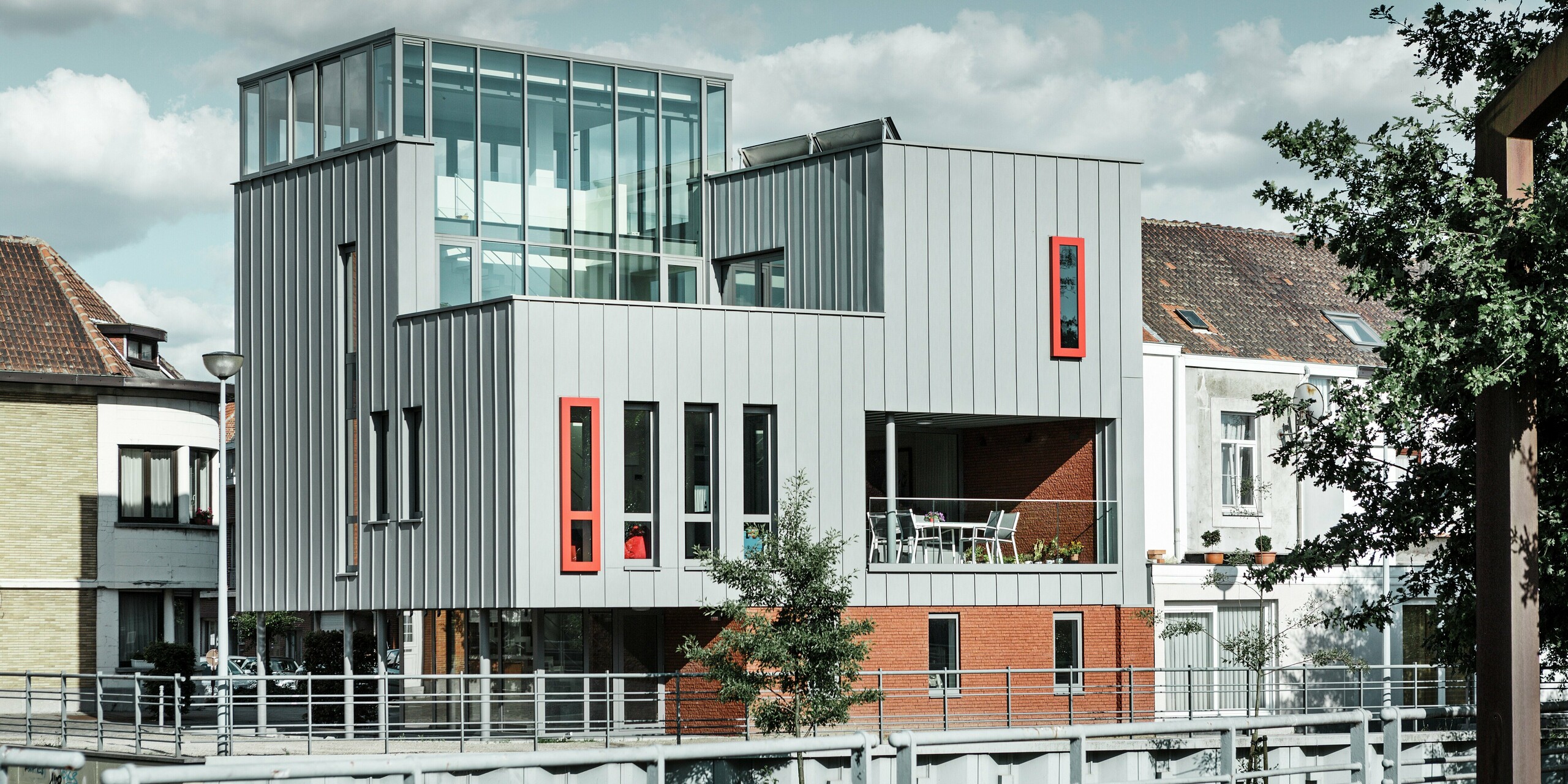 Moderne Architektur am Flussufer: Das Wohn-und Bürogebäude kombiniert eine elegante PREFALZ Fassade in Patinagrau mit auffälligen, roten Fensterrahmen als Eyecatcher. Die klar strukturierte Stehfalzoptik trifft auf großzügige Glasflächen und eine überdachte Terrasse. Aluminium als Baumaterial überzeugt hier durch Wetterbeständigkeit, Formstabilität und zeitloses Design.