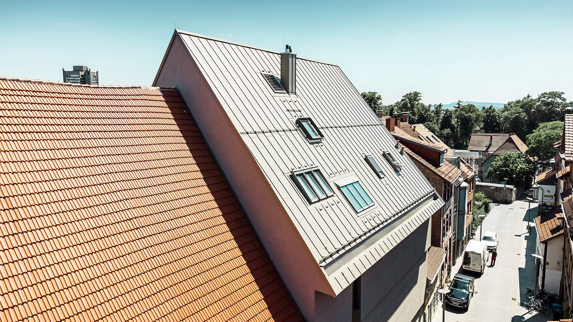 A side view of the exceptional roof; next to it, buildings covered with rusty red plain tiles.