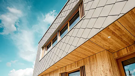 Die Nahaufnahme der Kindertagesstätte in Alzenau zeigt die präzise Ausführung und hochwertige Materialkombination aus bronzefarbenen PREFA Wandrauten 44 × 44 und naturbelassenem Holz. Die diagonale Struktur der Aluminiumfassade bildet ein markantes Schuppenmuster, das sich harmonisch mit den vertikalen Holzlamellen an den Fenstern und der fein ausgearbeiteten Holzuntersicht verbindet. Die Aufnahme aus der Froschperspektive betont die exakte Linienführung und handwerkliche Qualität der vorgehängten hinterlüfteten Fassade. Diese Detailansicht unterstreicht die gestalterische Stärke und Langlebigkeit von PREFA Produkten – ideal für nachhaltige Architekturprojekte, die Ästhetik, Funktionalität und Ökologie auf höchstem Niveau vereinen.