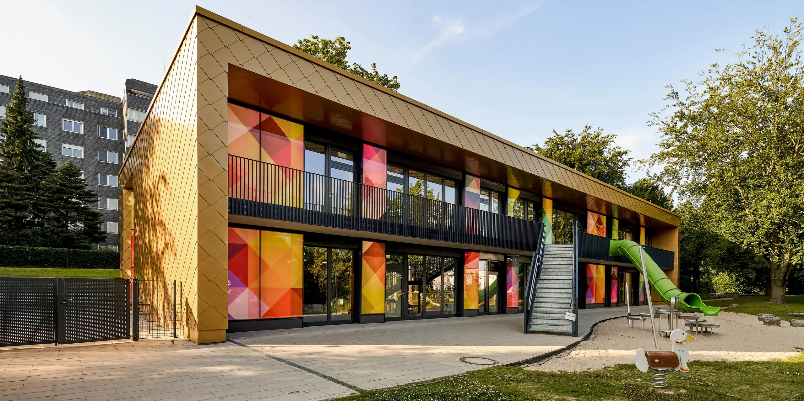 Hier trifft zeitgemäße Architektur auf eine spektakuläre PREFA Aluminiumfassade mit goldenen Wandrauten. Großflächige Verglasungen mit farbintensiven, geometrischen Elementen setzen lebendige Akzente und schaffen spannende Kontraste. Der offene Zugang sowie der Balkon und der Spielbereich unterstreichen die Nutzung als Kindertagesstätte. Die PREFA Aluminiumprodukte überzeugen durch extreme Langlebigkeit, Witterungsbeständigkeit und brillante Farbwirkung. Sie sind eine wartungsfreie, nachhaltige Lösung für ausdrucksstarke Fassaden mit hoher Designqualität.