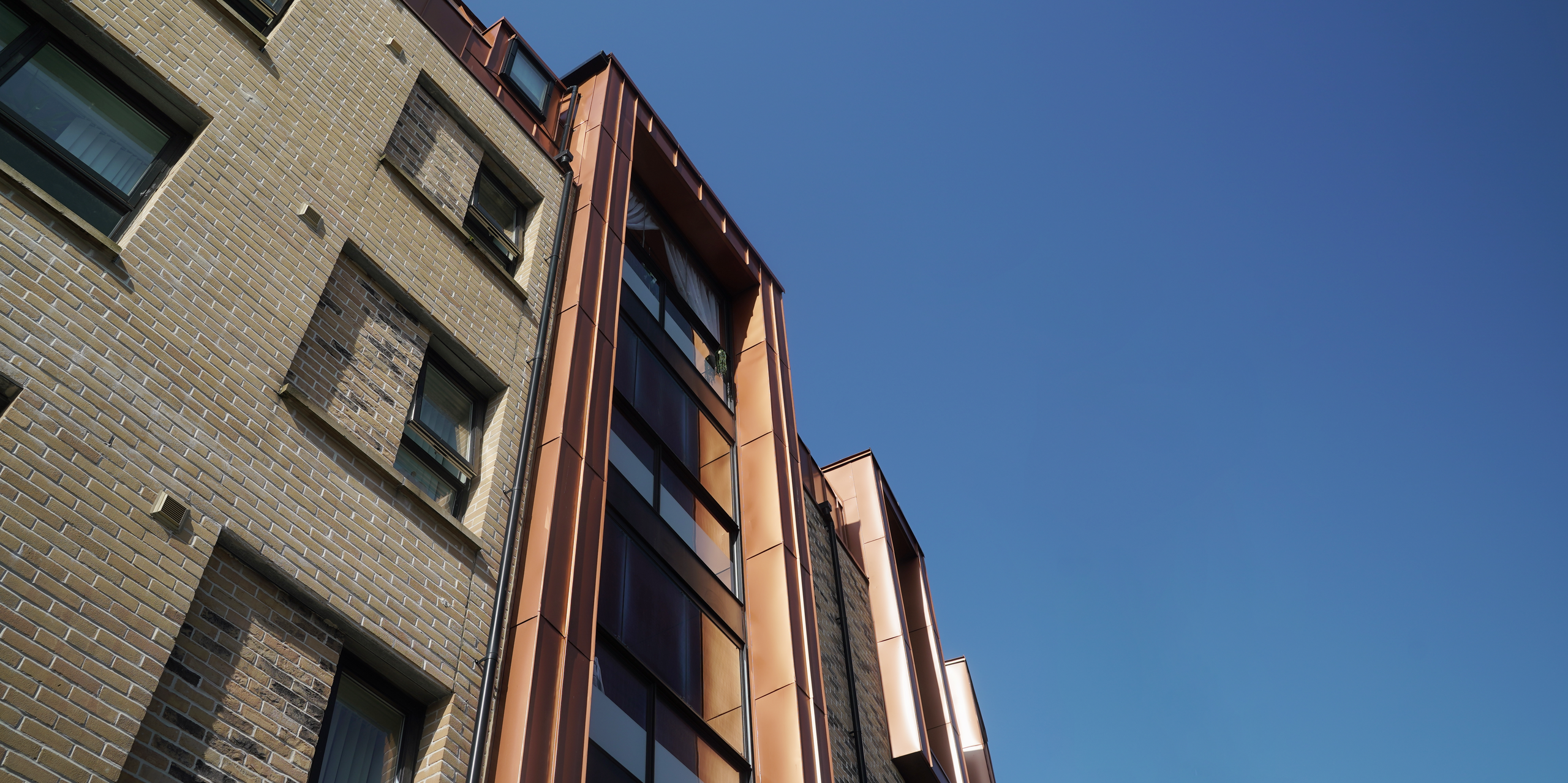 Perspective view of the façade of a modern residential development on Nethan Street in Glasgow, clad with FALZONAL in New Copper. The metallic façade elements catch and reflect the light, emphasising the shape of the building and providing an impressive visual contrast to the traditional brick architecture. The PREFA aluminium product not only provides an appealing aesthetic, but also promises durability and low maintenance.