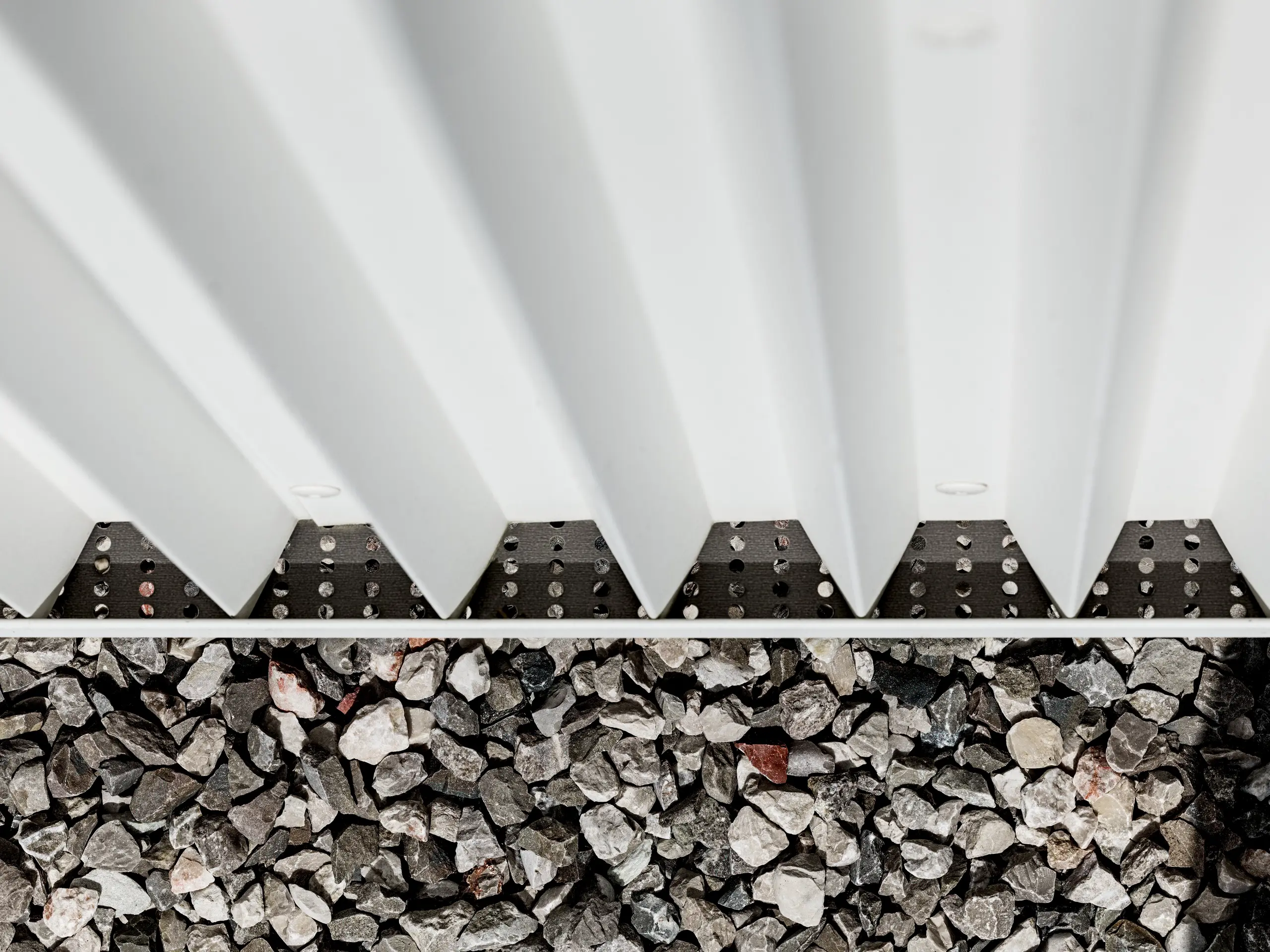 Das Bild zeigt einen Detailausschnitt der hinterlüfteten PREFALZ-Fassade aus reinweißem Aluminium, die mit präzise gefalteten Zickzack-Formen versehen ist. Diese innovative Falztechnik gewährleistet nicht nur eine optisch ansprechende Struktur, sondern auch eine hervorragende Belüftung und Witterungsbeständigkeit der Fassade. Die Verbindung der Fassadenelemente ist durch spezialisierte, unsichtbare Befestigungen realisiert, die eine besonders saubere und langlebige Oberfläche ermöglichen. Der untere Bereich des Bildes zeigt den Kiesbelag, der in der Umgebung des Gebäudes zur Drainage und Stabilisierung dient, was die Funktionalität der Fassadengestaltung weiter unterstreicht. Durch die Verwendung von PREFALZ werden ästhetische Ansprüche und technische Anforderungen gleichermaßen erfüllt, was das Material zu einer idealen Wahl für moderne und nachhaltige Architektur macht.