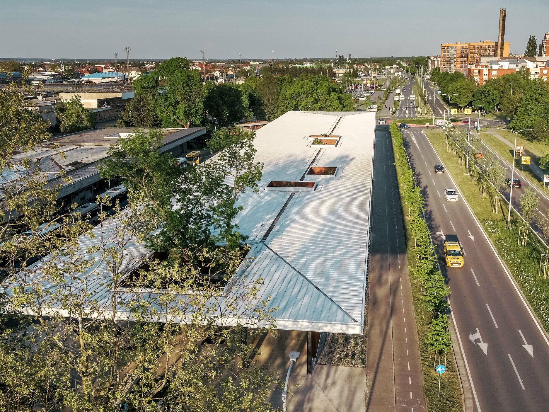 The picture shows an expansive view of a modern bus stop in Croatia, covered with a white Prefalz roof from PREFA. The roof is interrupted by several rectangular openings from which trees protrude. The bus stop is located next to a wide, two-lane road lined with trees. Various buildings and residential houses that characterise the urban surroundings can be seen in the background.