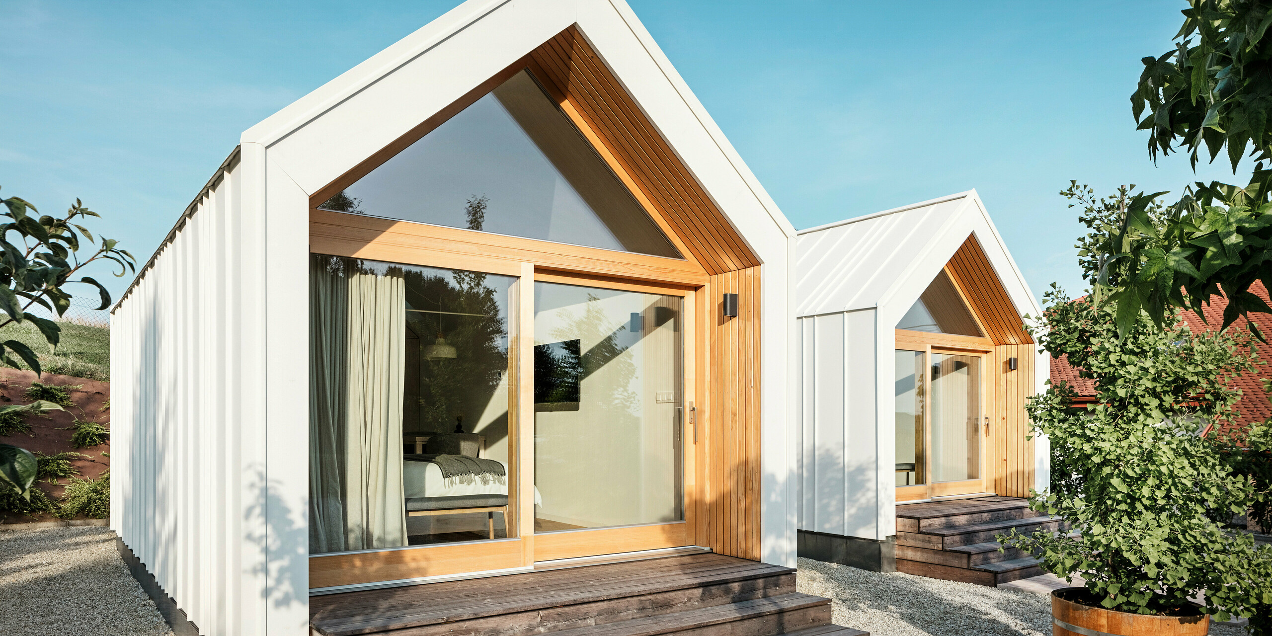 Front view of the two tiny houses of the 'Pri Momi' guest accommodation in Kančevci, Slovenia, with large glass fronts, designed with PREFALZ roof and façade cladding in P.10 pure white and natural wood. The minimalist micro houses have wide wooden steps leading up to the entrances and offer a direct view into the cosy interiors. The clear lines and the combination of aluminium and wood underline the modern and inviting design of the holiday accommodation.