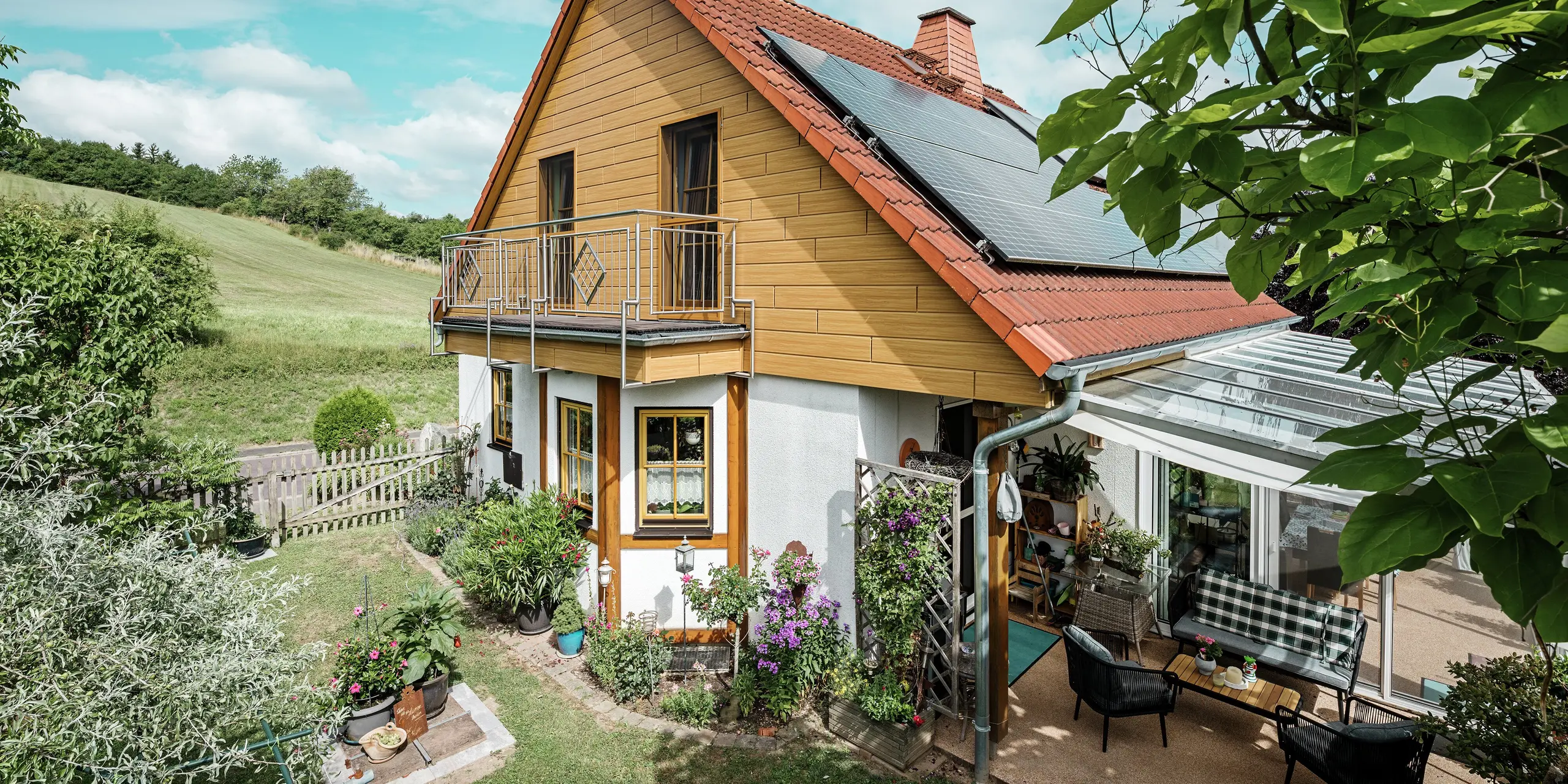 This image shows a charming detached house in Blieskastel whose freshly renovated gable with PREFA natural oak sidings is immediately striking. The warm wood look of durable aluminium harmonises impressively with the red tiled roof and the spacious conservatory. An idyllic, vibrant scene is created, surrounded by lush greenery and lovingly-planted flower beds. The robust façade renovation with PREFA not only gives the house a natural look but is also impressive in terms of its weather resistance, colour fidelity and virtually maintenance-free durability.