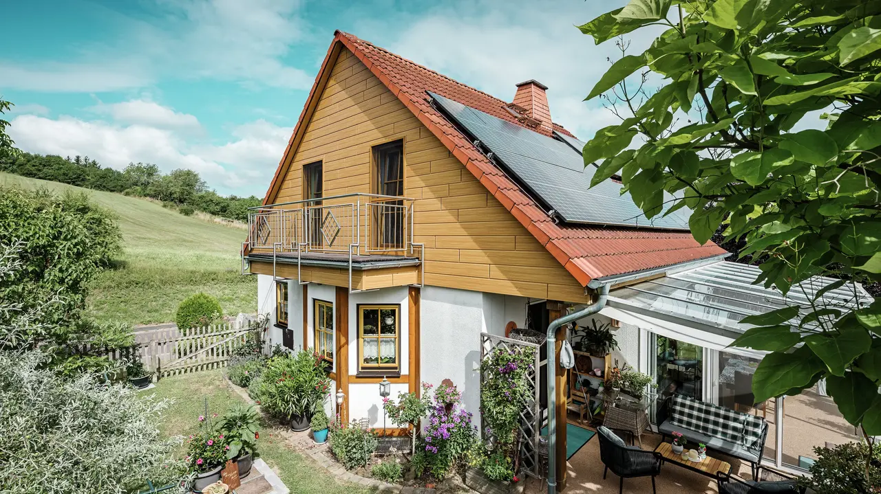 This image shows a charming detached house in Blieskastel whose freshly renovated gable with PREFA natural oak sidings is immediately striking. The warm wood look of durable aluminium harmonises impressively with the red tiled roof and the spacious conservatory. An idyllic, vibrant scene is created, surrounded by lush greenery and lovingly-planted flower beds. The robust façade renovation with PREFA not only gives the house a natural look but is also impressive in terms of its weather resistance, colour fidelity and virtually maintenance-free durability.