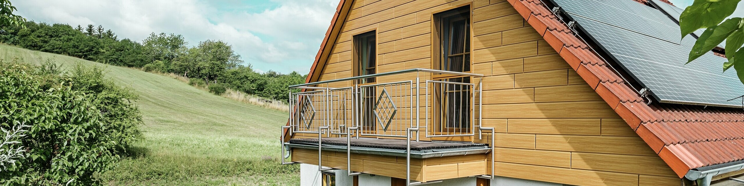 This image shows a charming detached house in Blieskastel whose freshly renovated gable with PREFA natural oak sidings is immediately striking. The warm wood look of durable aluminium harmonises impressively with the red tiled roof and the spacious conservatory. An idyllic, vibrant scene is created, surrounded by lush greenery and lovingly-planted flower beds. The robust façade renovation with PREFA not only gives the house a natural look but is also impressive in terms of its weather resistance, colour fidelity and virtually maintenance-free durability.
