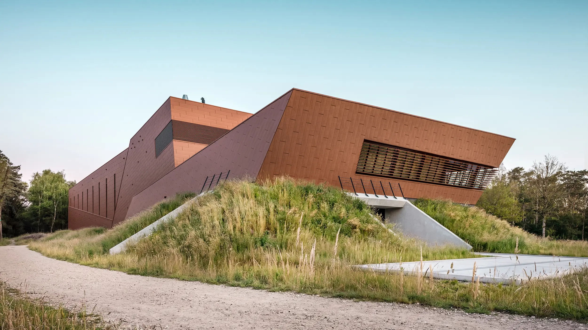 Architektonisch expressives Trinkwasserpumpwerk WML in Lottum mit skulptural gefalteten Baukörpern, eingebettet in eine naturnahe Böschungslandschaft. Die markante PREFA Siding.X Aluminiumfassade in warmer Rostoptik zieht mit ihrer facettierten, lebendigen Oberfläche alle Blicke auf sich und spielt eindrucksvoll mit Licht und Schatten. Horizontale Lamellen und präzise Linienführung betonen die dynamische Geometrie. Die leichten, langlebigen und vollständig recycelbaren PREFA Aluminium-Paneele stehen für höchste Witterungsbeständigkeit, nachhaltige Bauweise und eine wartungsarme Gebäudehülle.